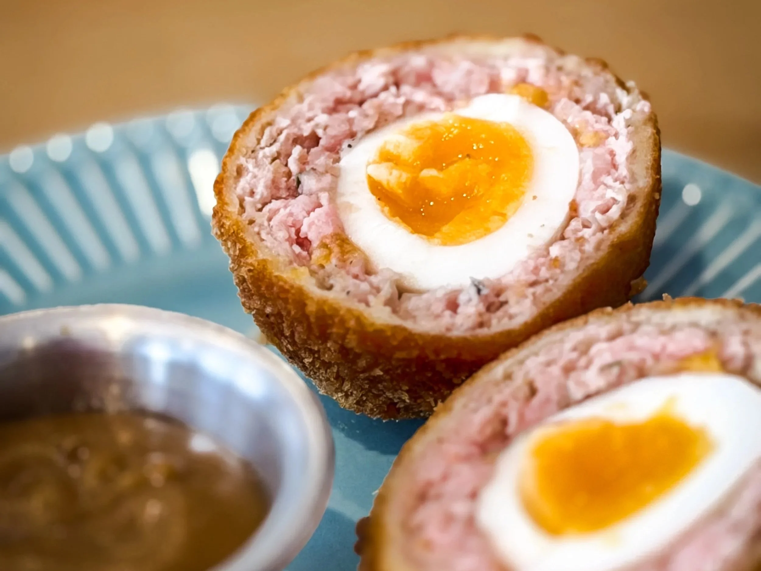 Close-up of a halved Scotch egg showing a soft-boiled egg yolk and pink sausage meat enclosed in a breaded outer layer, placed on a blue plate.