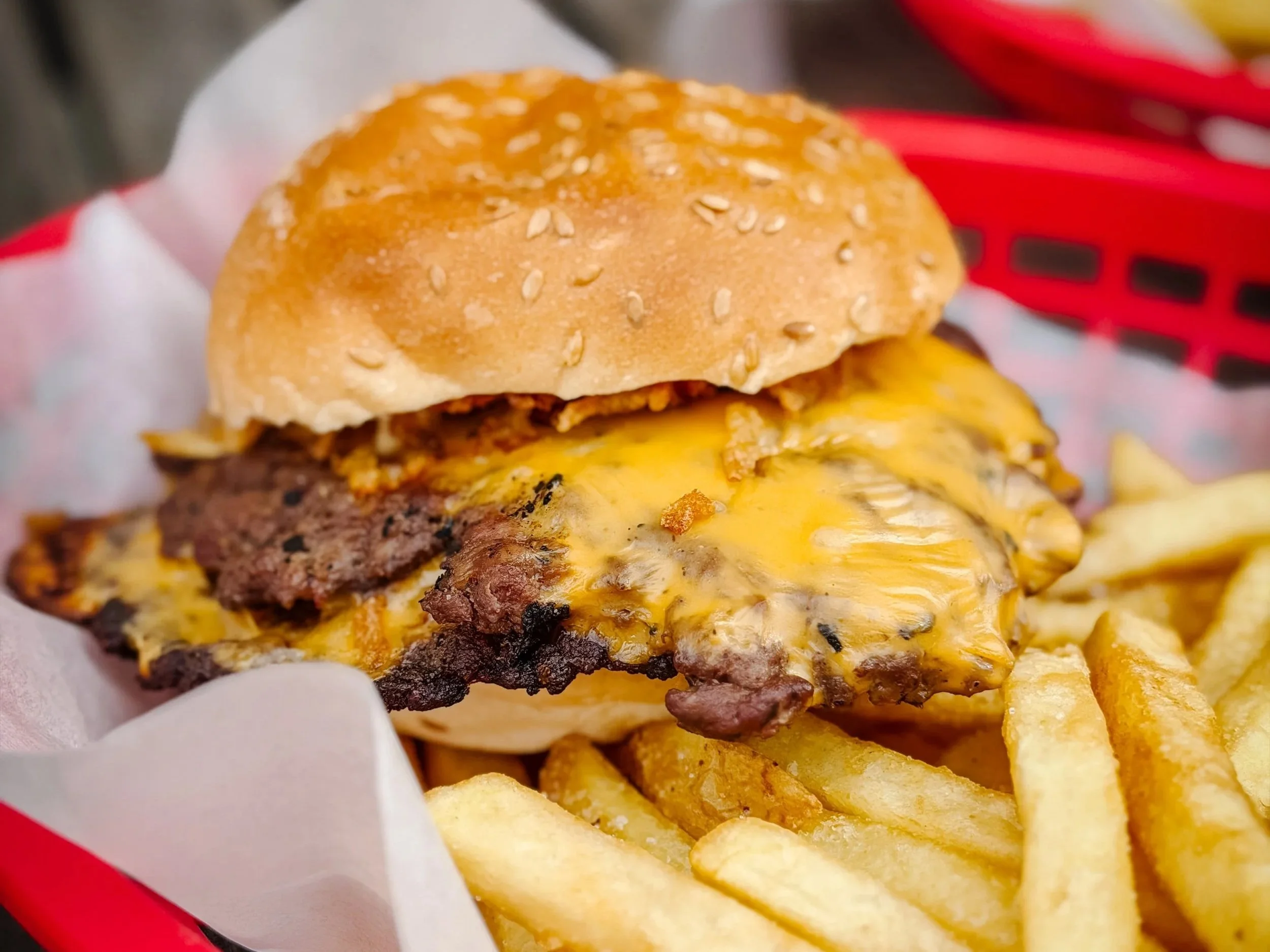 A close-up of a cheeseburger with a sesame seed bun, melted cheese, a beef patty, and crispy potato chips in the background, in a red basket lined with white paper.