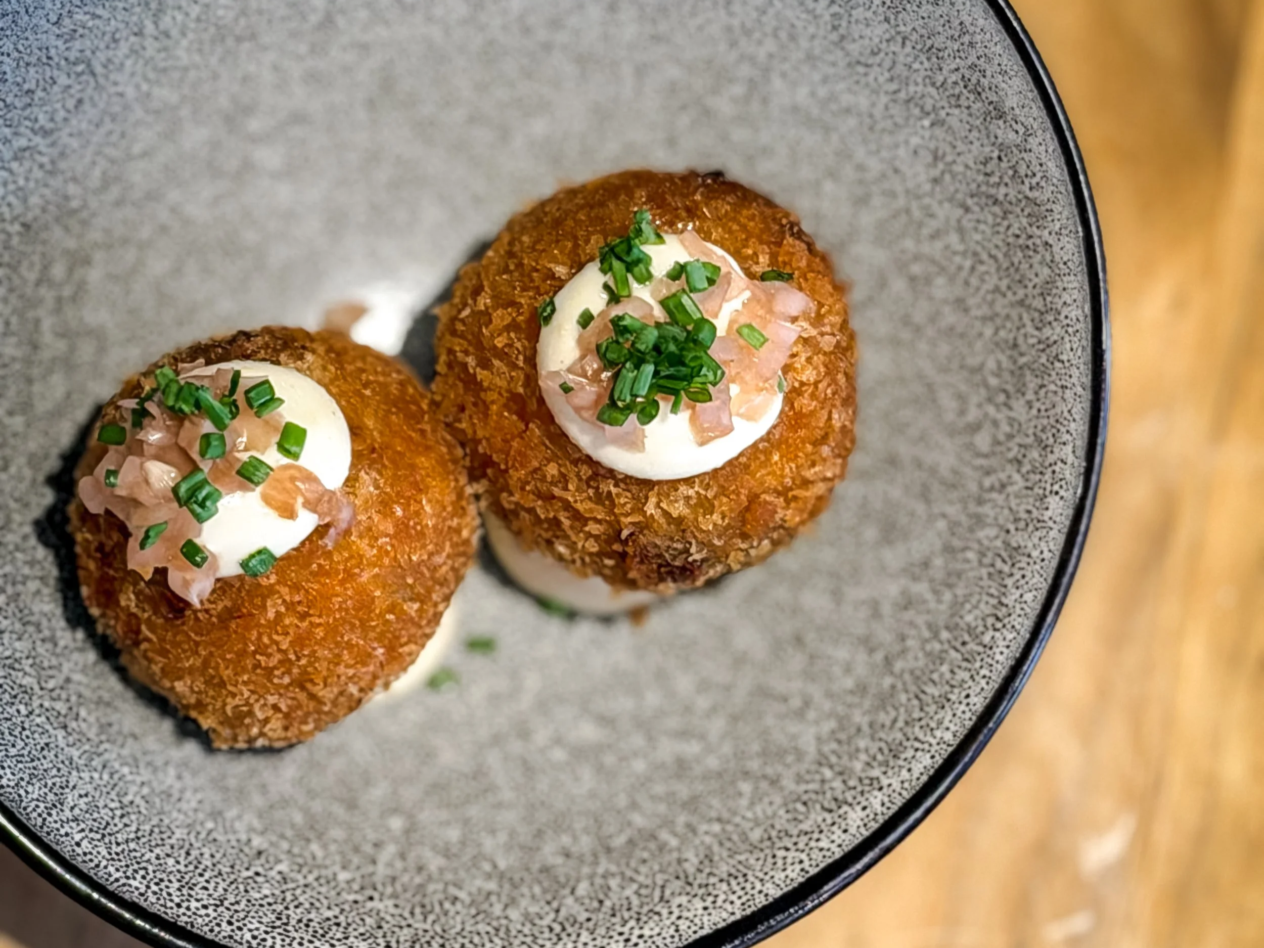 Close-up of three fried croquettes topped with sour cream, chopped ham, and chopped green onions on a gray textured plate.