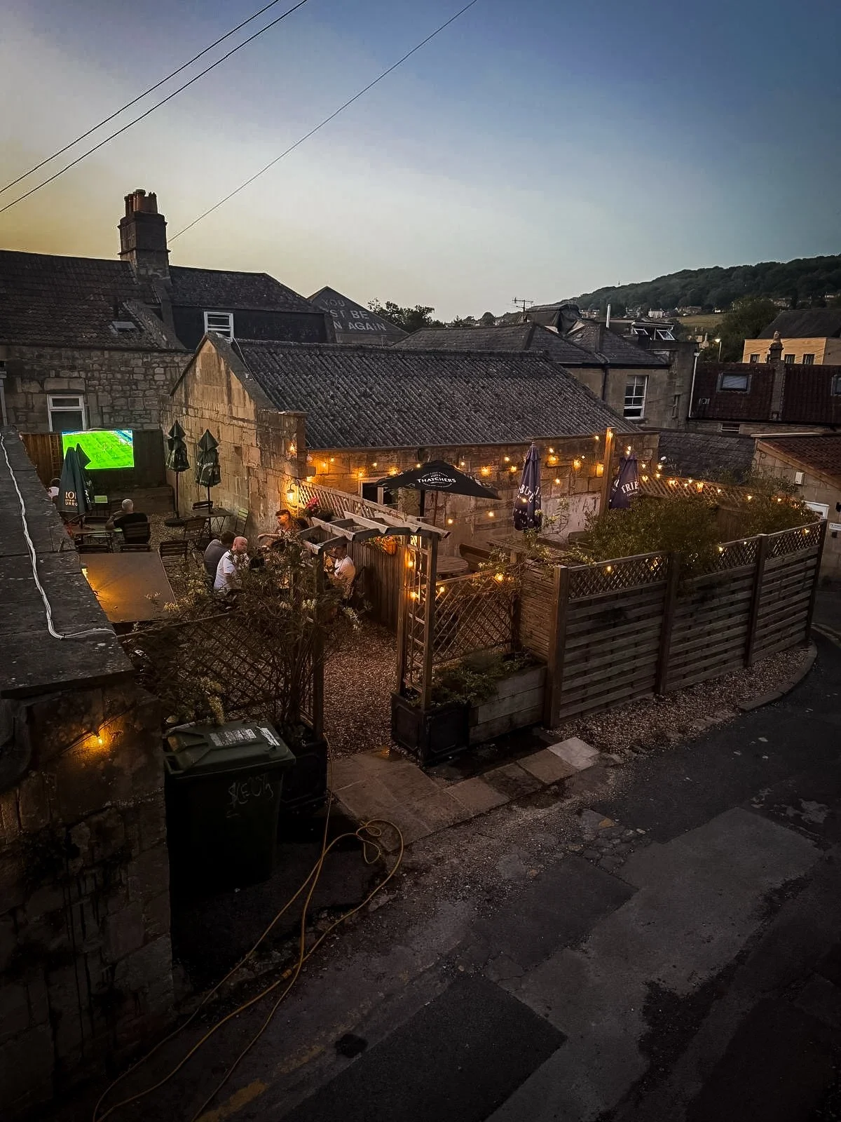 An outdoor patio area at twilight with string lights, tables, and umbrellas. Several people are sitting and chatting, and there is a fence surrounding the patio.