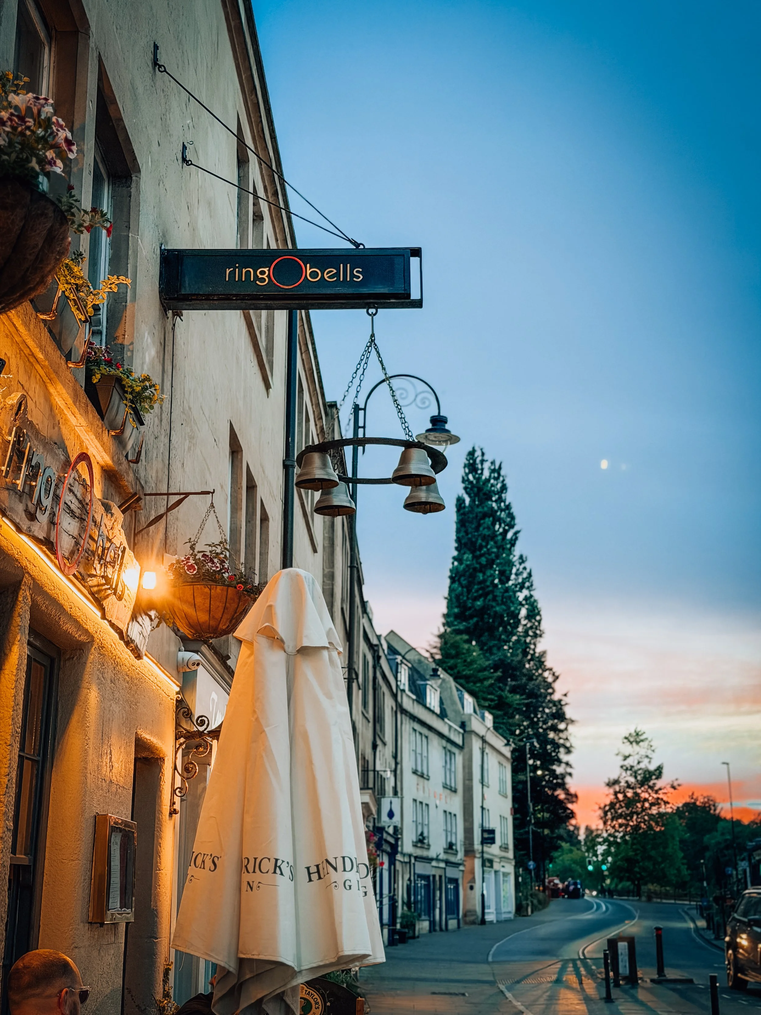 A street scene at dusk with a building on the left featuring hanging flower pots, a sign for 'ringo bells,' and outdoor seating with umbrellas. The street curves to the right with trees and a small hill in the background, and the sky is a mix of blue and pink with the moon visible.