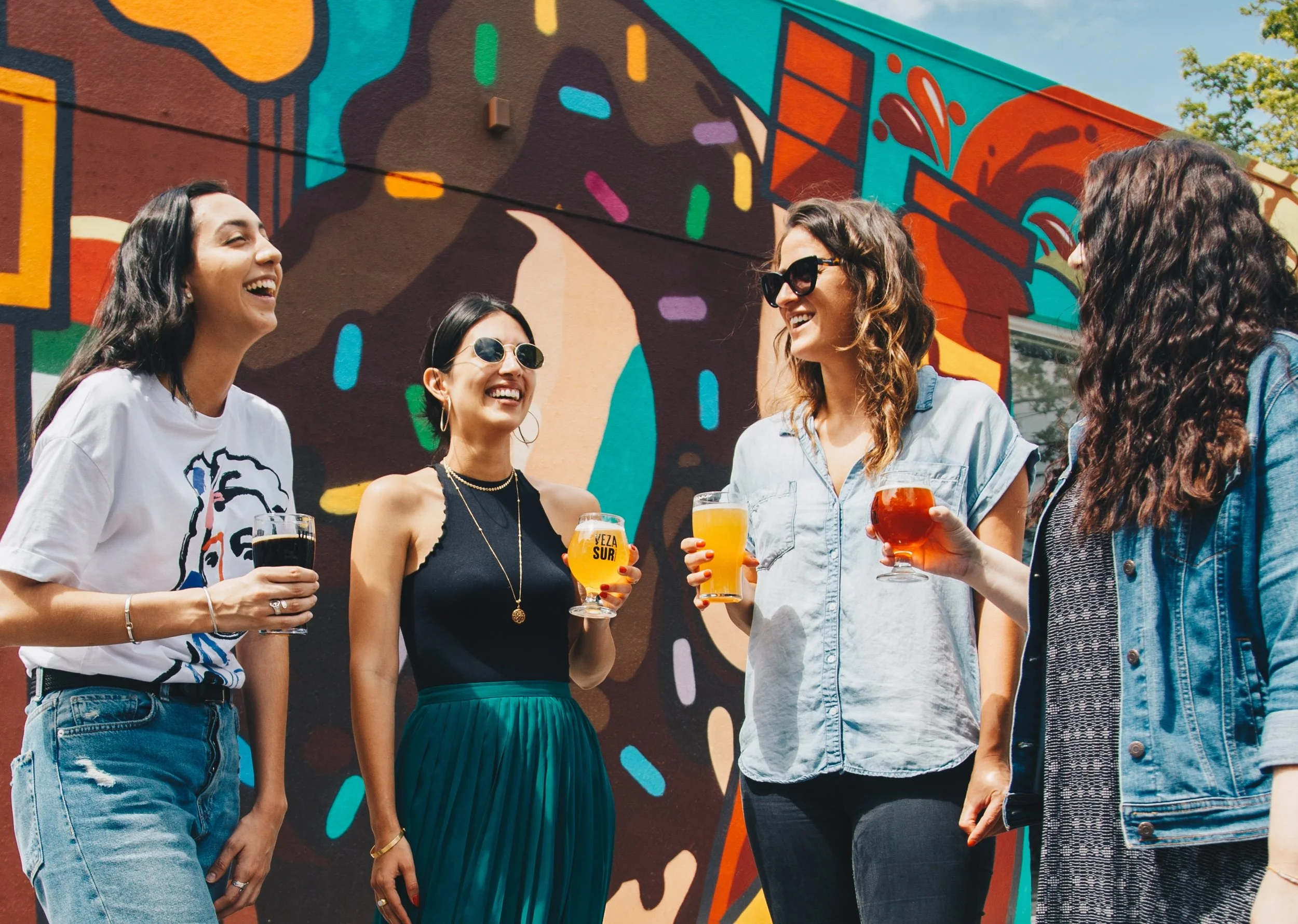 Group of five women standing outdoors, holding drinks, chatting and smiling, with a colorful mural on the wall behind them.