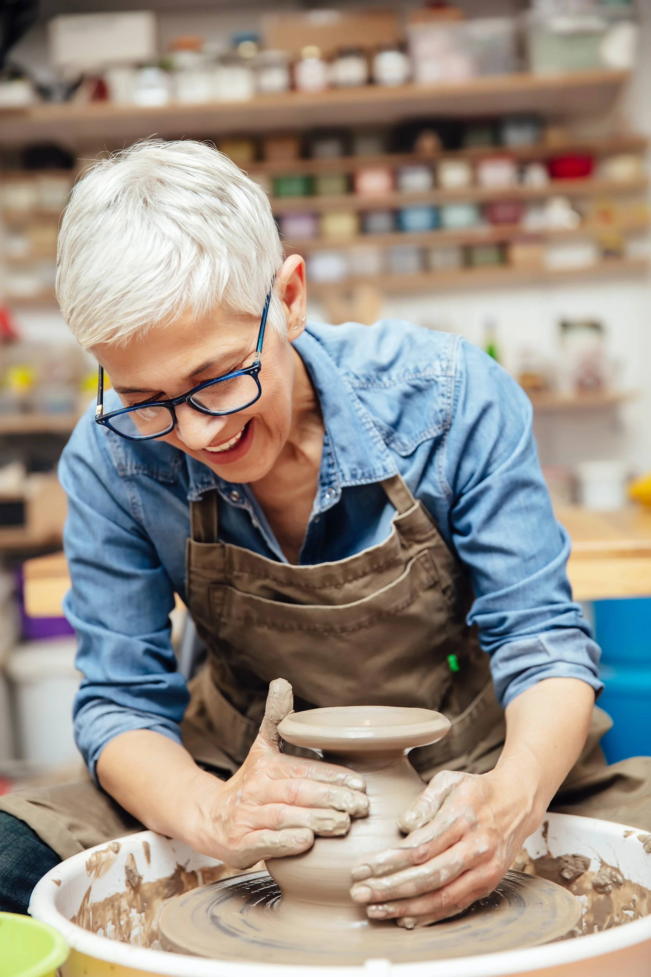 Older woman with short white hair and glasses creating a pottery piece on a pottery wheel in a ceramics studio.