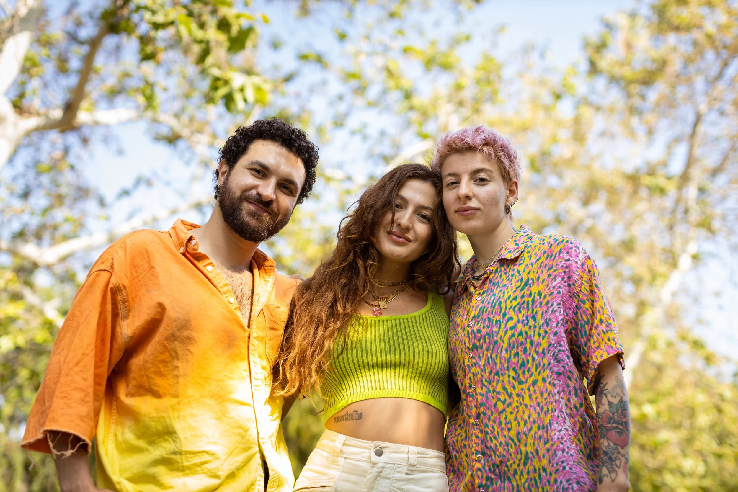 Group of three young adults standing outdoors, smiling at the camera, with trees and blue sky in the background.