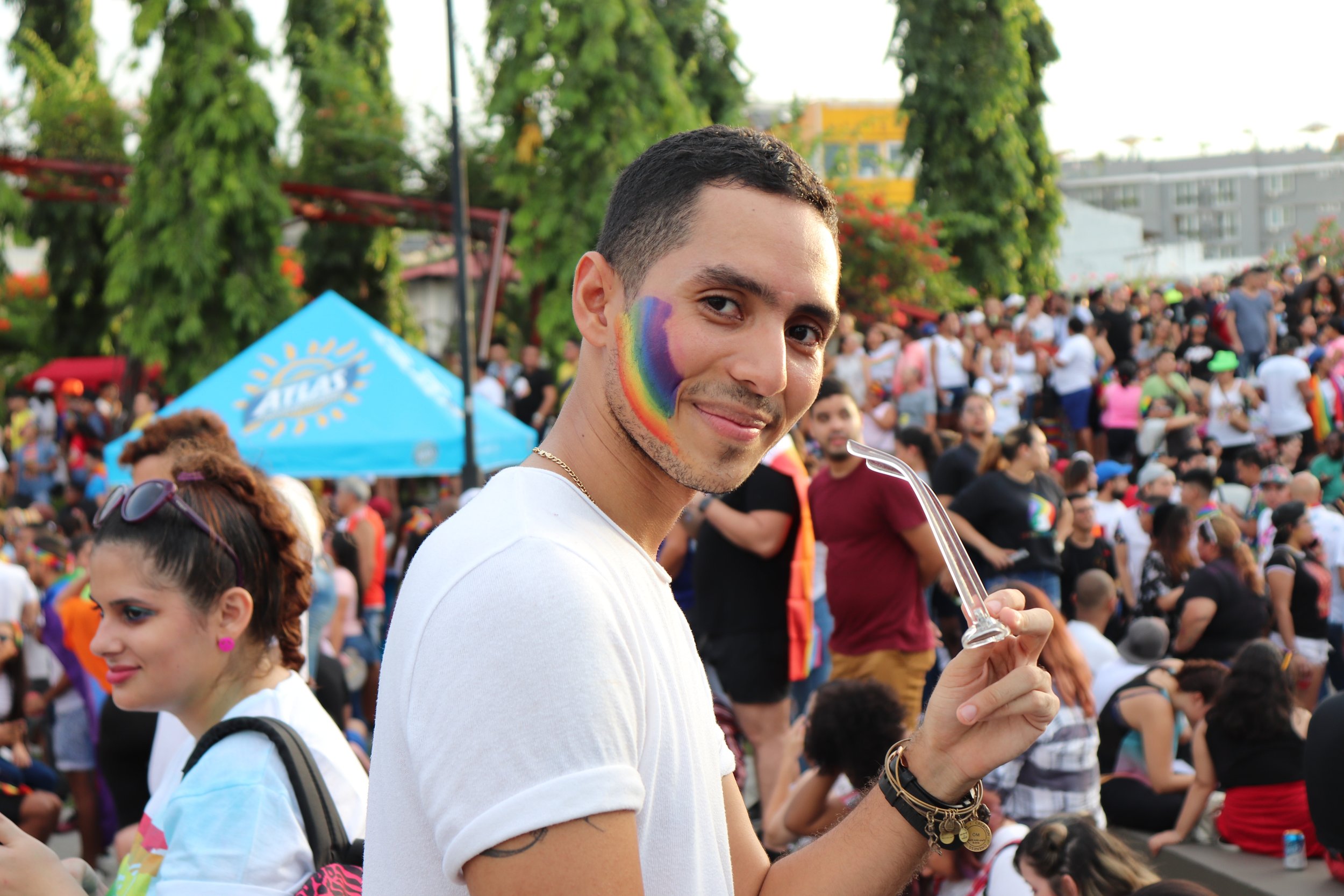 A young man smiling at a pride celebration, with rainbow face paint on his cheek and holding a glass container, surrounded by a diverse crowd of people, some with rainbow accessories, at an outdoor event with trees and a blue tent in the background.