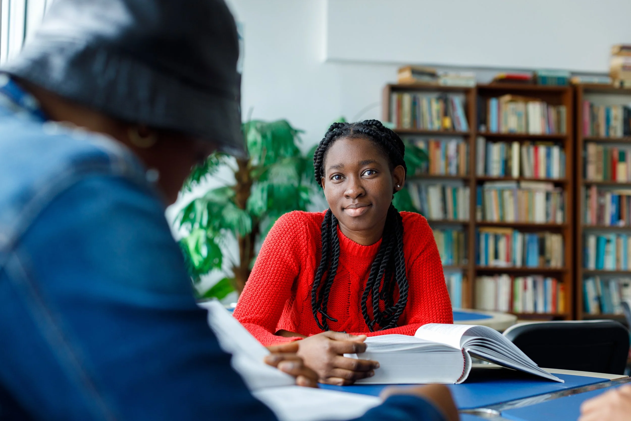 Young woman with braided hair wearing a red sweater sitting at a table in a library, engaged in conversation with a person whose face is not visible.