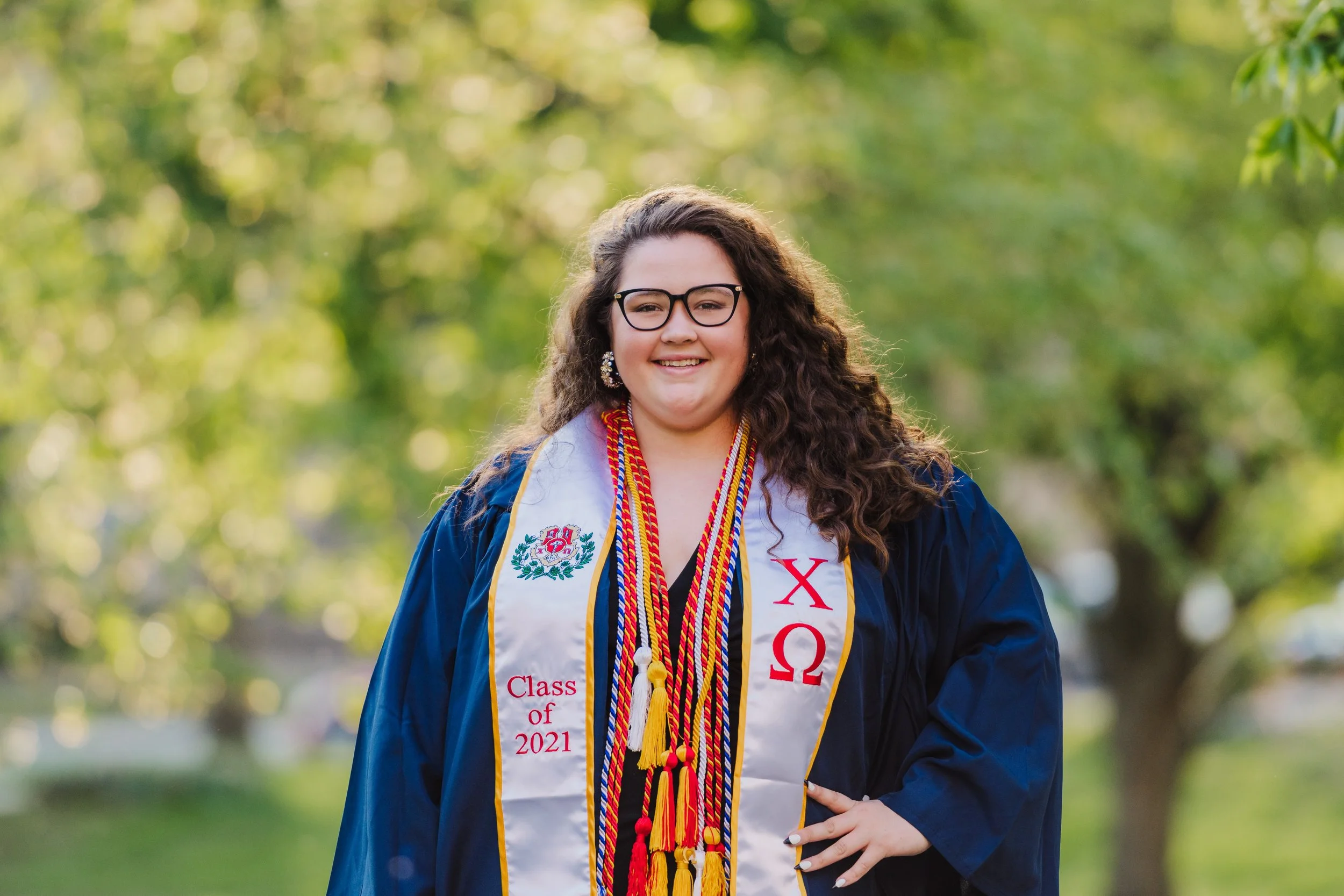 A young woman with curly hair wearing glasses and a graduation gown, smiling at the camera, outdoors with green trees in the background, decorated with honor cords, stoles, and a sash.