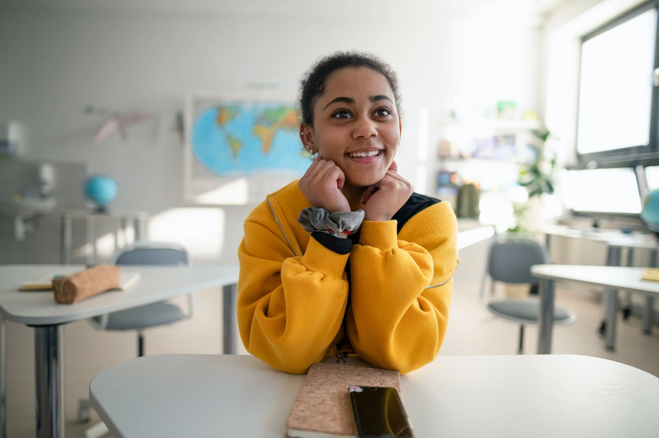 A young girl with curly hair, wearing a yellow sweatshirt, smiling and resting her chin on her hands at a desk in a classroom, with a world map on the wall in the background.