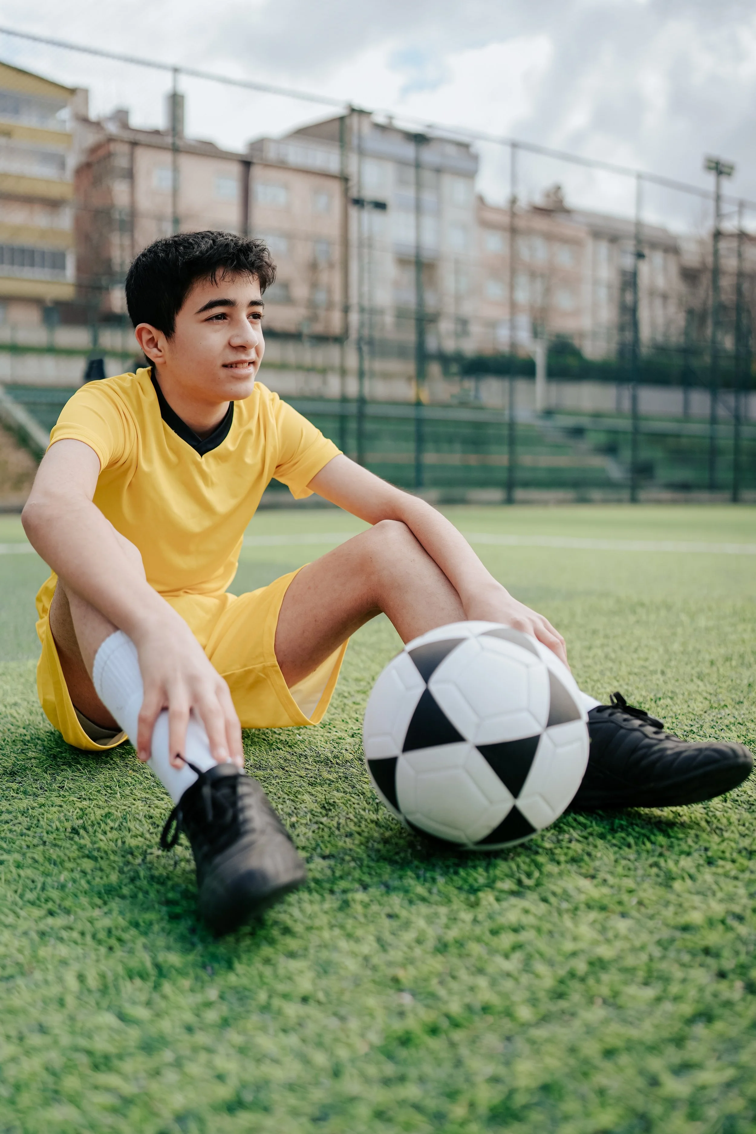 A young male soccer player wearing a yellow kit sitting on the field, stretching with a soccer ball in front of him, at an outdoor sports facility with buildings and a cloudy sky in the background.
