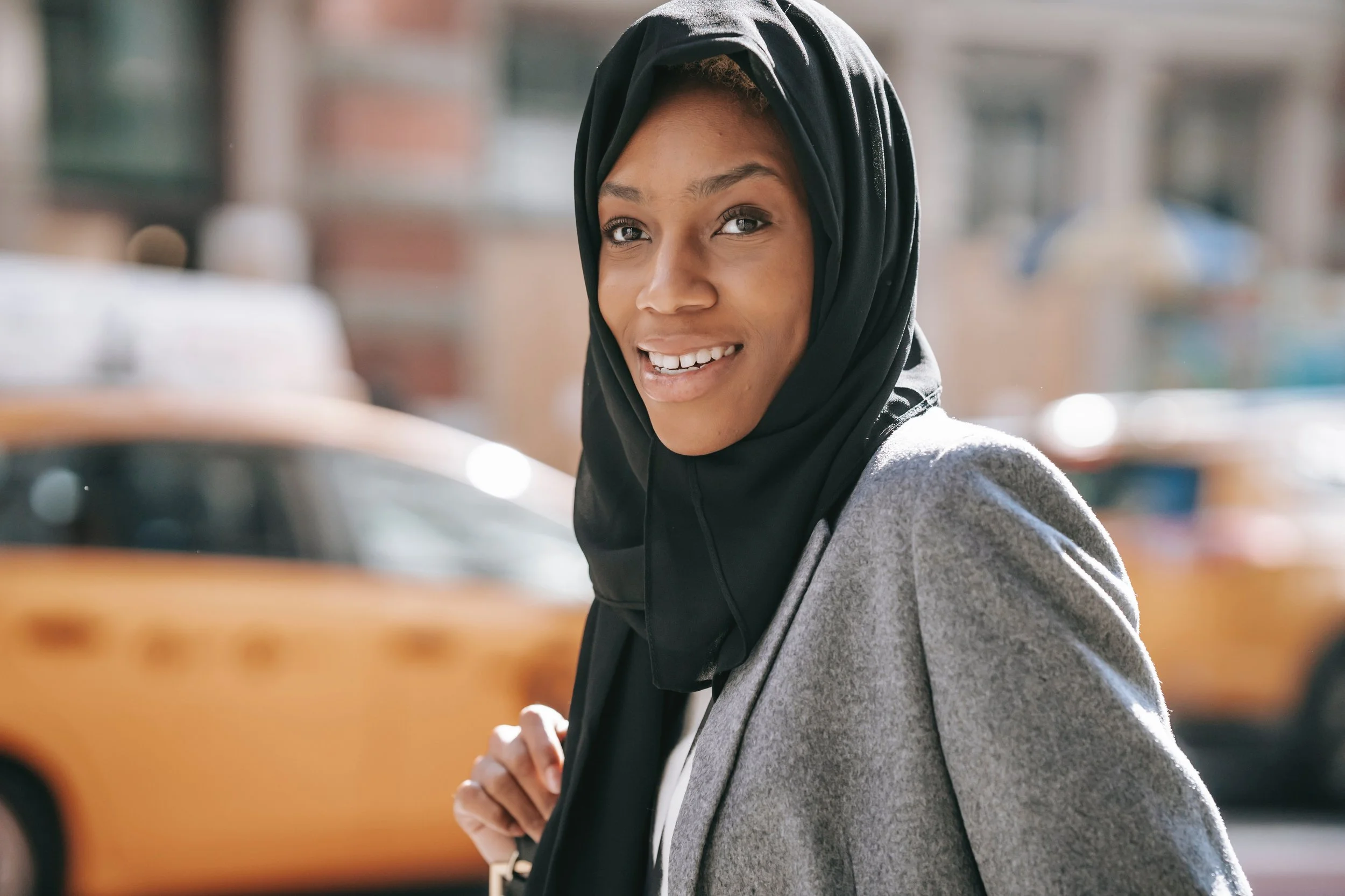 Young woman wearing a black headscarf and grey coat smiling outdoors with blurred background.