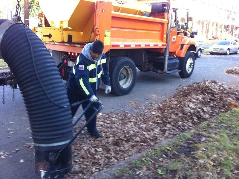 City worker raking leaves into a pile on the sidewalk next to a street where a large orange maintenance truck is parked.