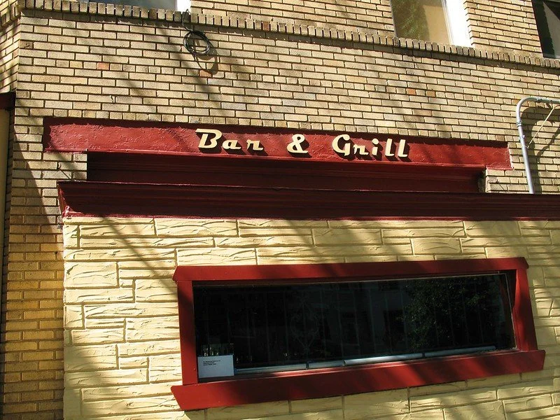 Exterior of a building with a sign that reads 'Bar & Grill' above a large window with a red frame.