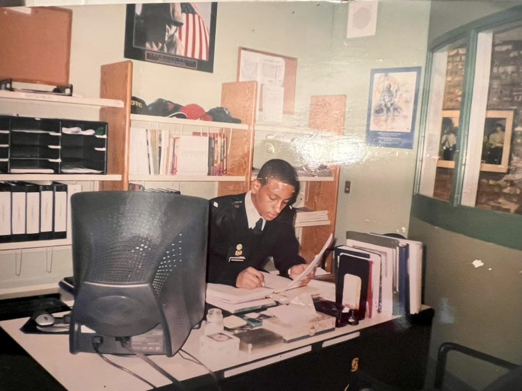 A uniformed woman working at a cluttered office desk with books, papers, and office supplies in a small, well-lit office.