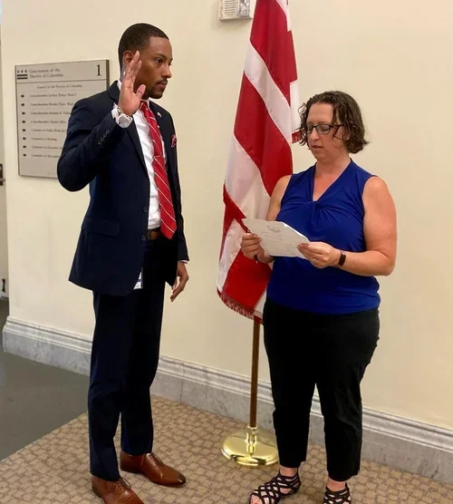 A woman holding a document and taking an oath during a swearing-in ceremony with a man in formal attire raising her right hand. The American flag is in the background.