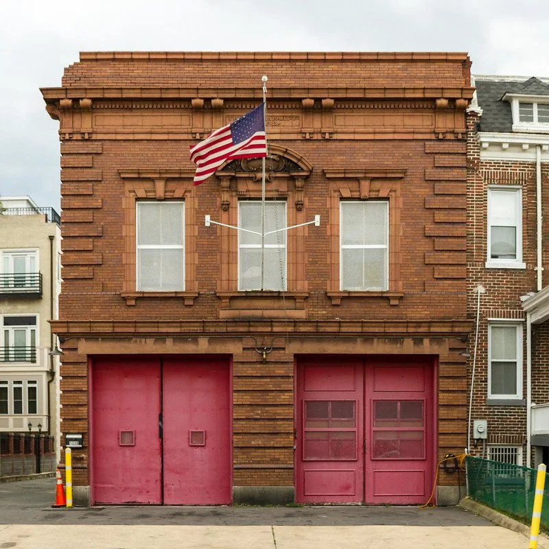 Two large red garage doors on a brick building, with two windows and an American flag flying above.