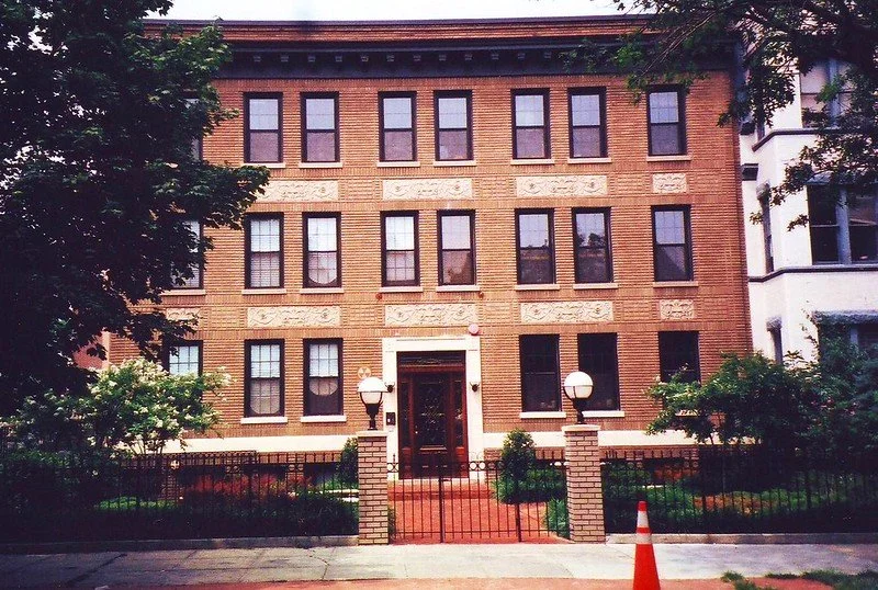 A brick multi-story residential building with a decorative facade, six windows on each floor, and a centered front door. The building has three brick columns with spherical lamps on top, a small front yard with plants, and is enclosed by a black fence. There is a traffic cone in the sidewalk in front.