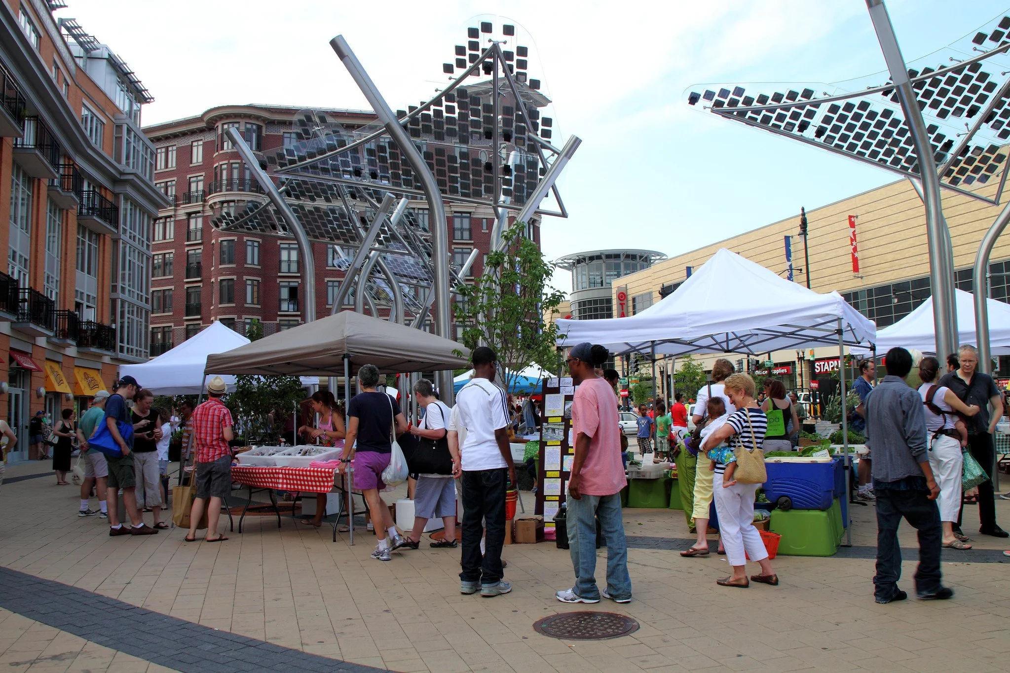 Crowd of people at an outdoor market with white tents and vendors selling produce, in a city setting with tall buildings and a blue sky.