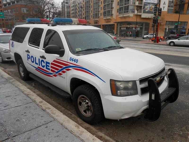 White police SUV with light bar on roof, police markings, bumper guard, parked on city street during rainy weather.