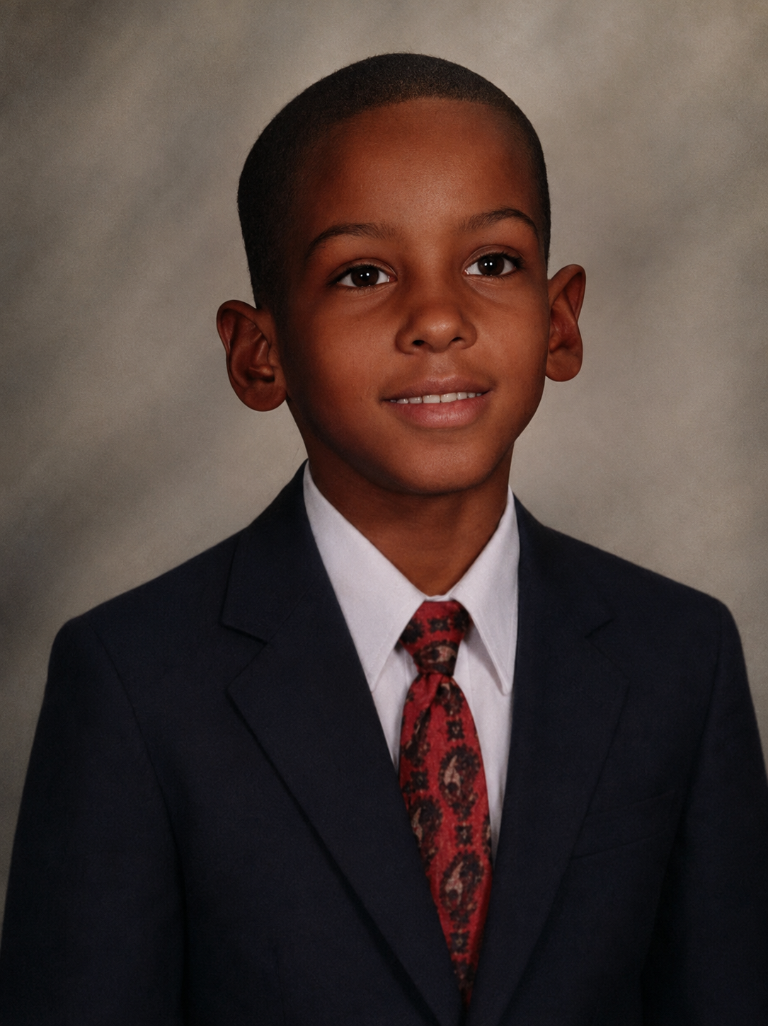 Young boy in formal suit with a white shirt and patterned tie, smiling for a portrait against a gray background.