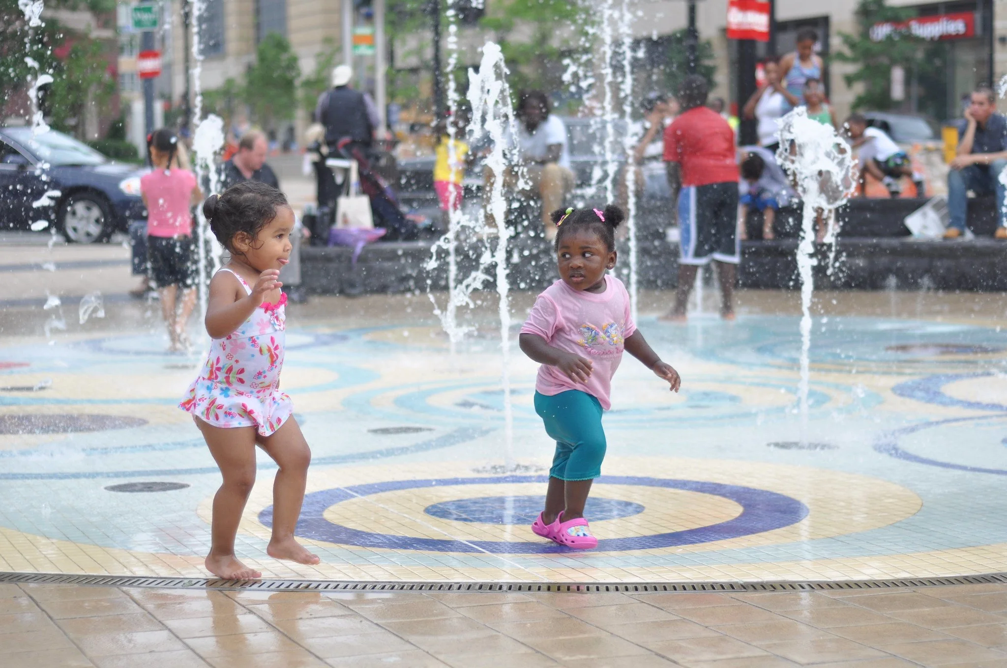 Two young girls playing in a water fountain in an urban park on a rainy day, with people in the background.