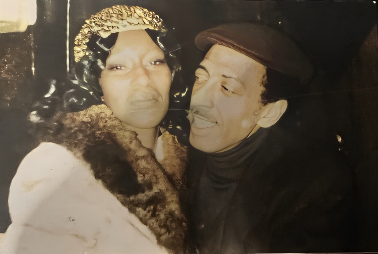 A black and white photo showing a man and woman close together, both wearing vintage clothing and hats, smiling and looking at each other.