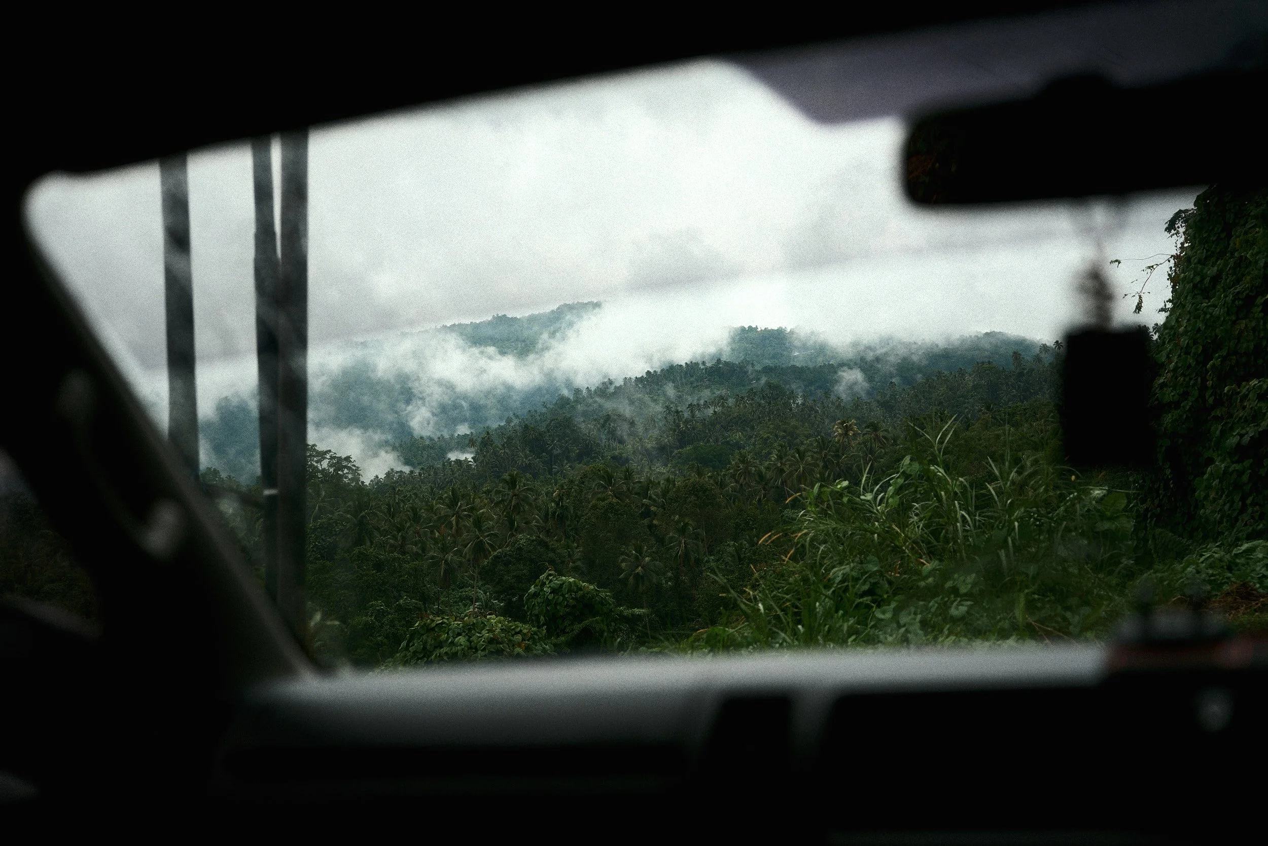 A view of lush, green mountains and trees through a vehicle window with clouds or fog surrounding the peaks.