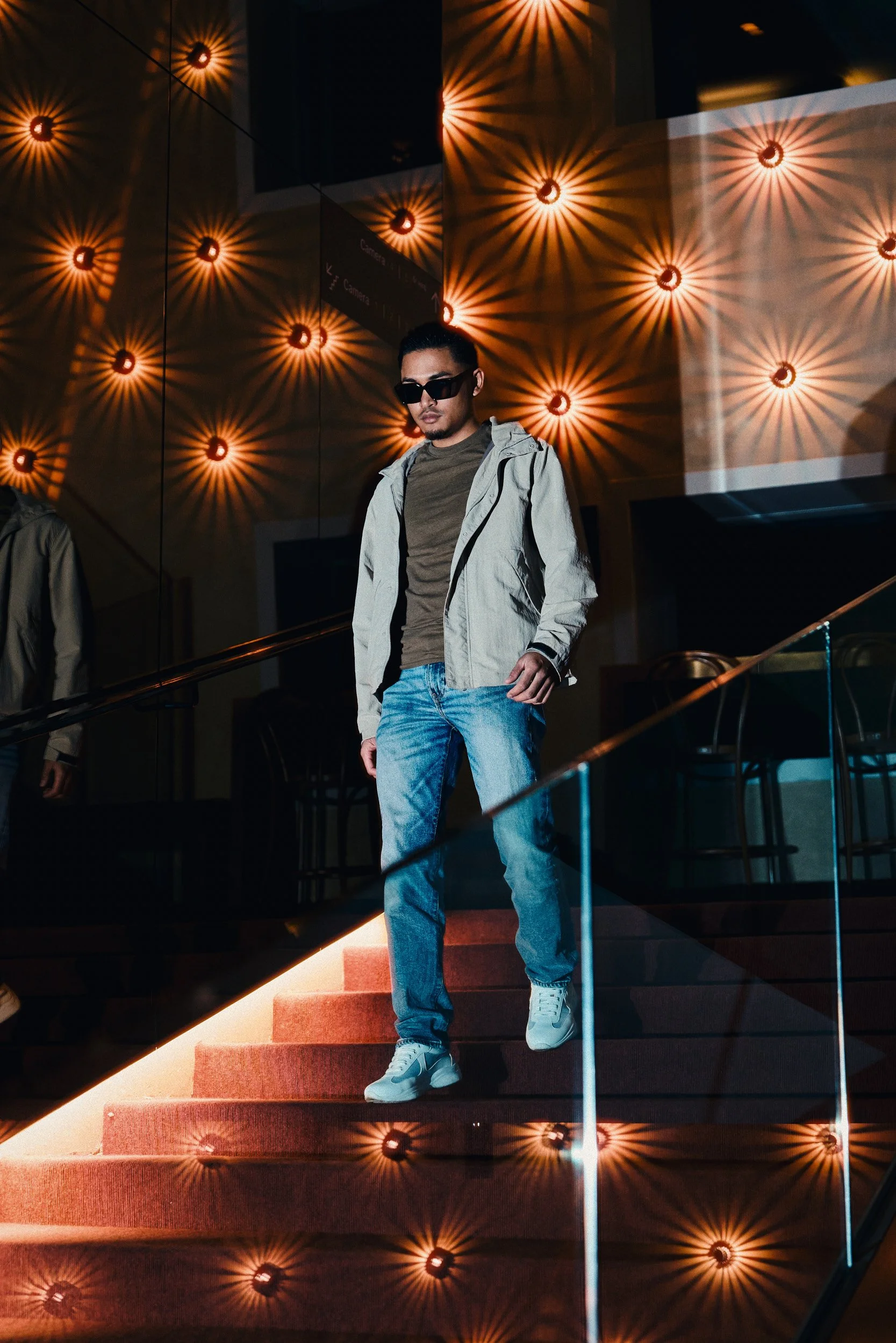 A young man in sunglasses, a gray jacket, a brown shirt, and blue jeans descending red-carpeted stairs inside a dimly lit building with decorative starburst wall lights.