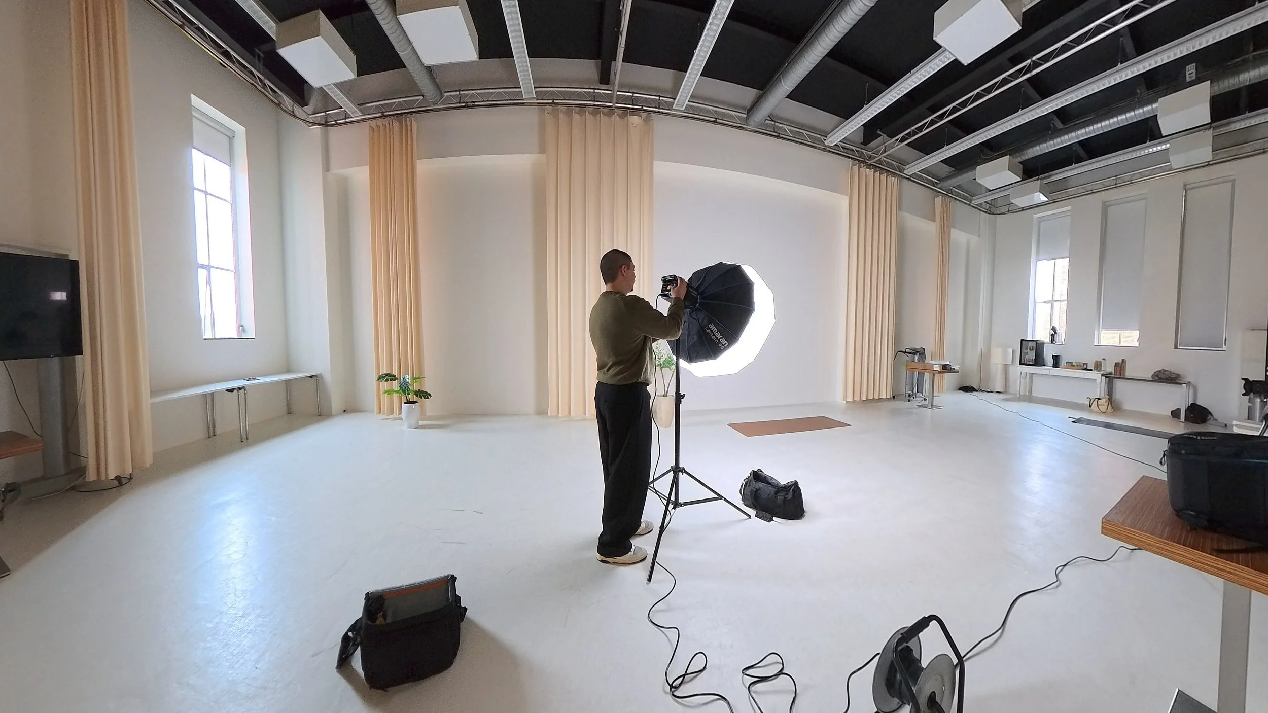 Videographer setting up in a spacious studio with large windows, beige curtains, and video equipment including a softbox light.