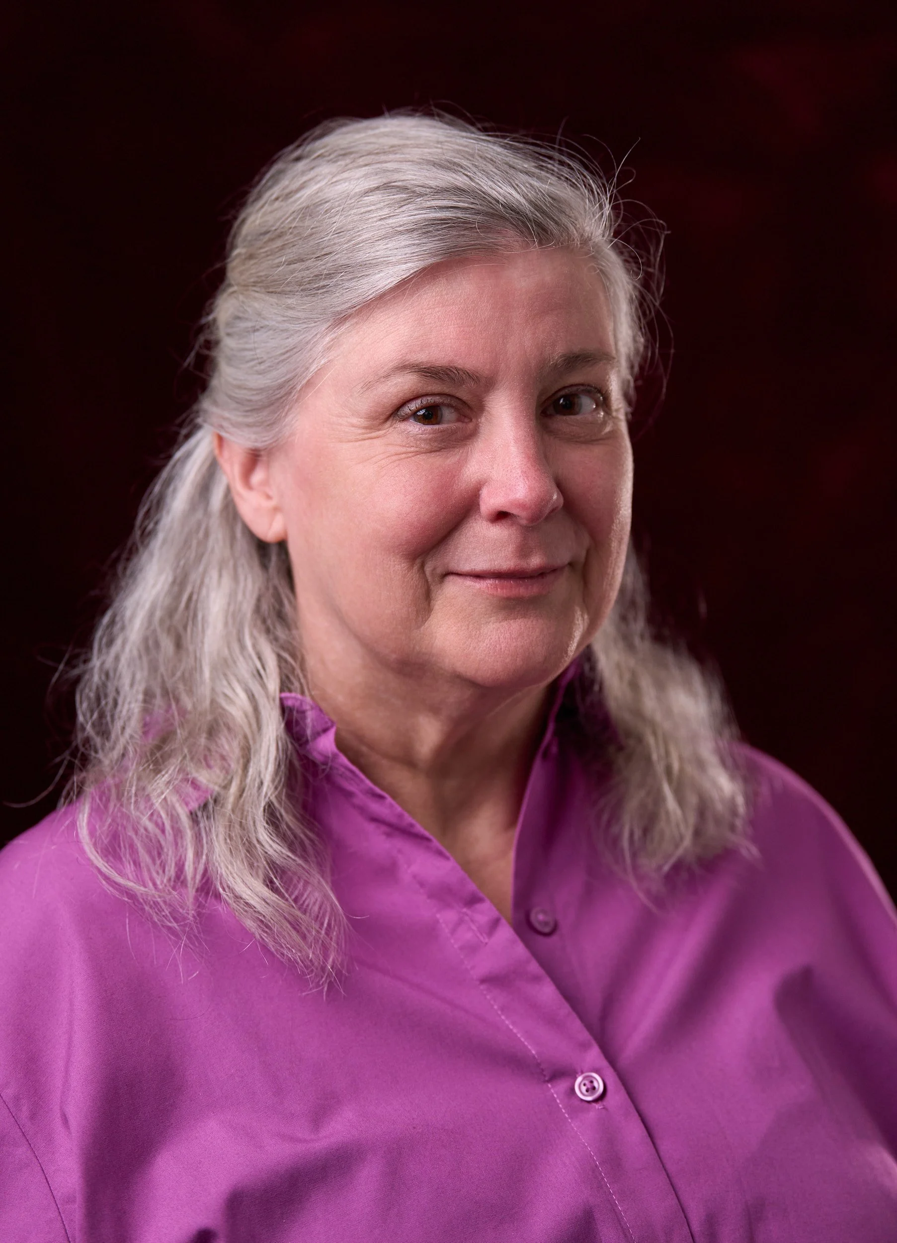 Close-up of a smiling older woman with long gray hair wearing a purple shirt.