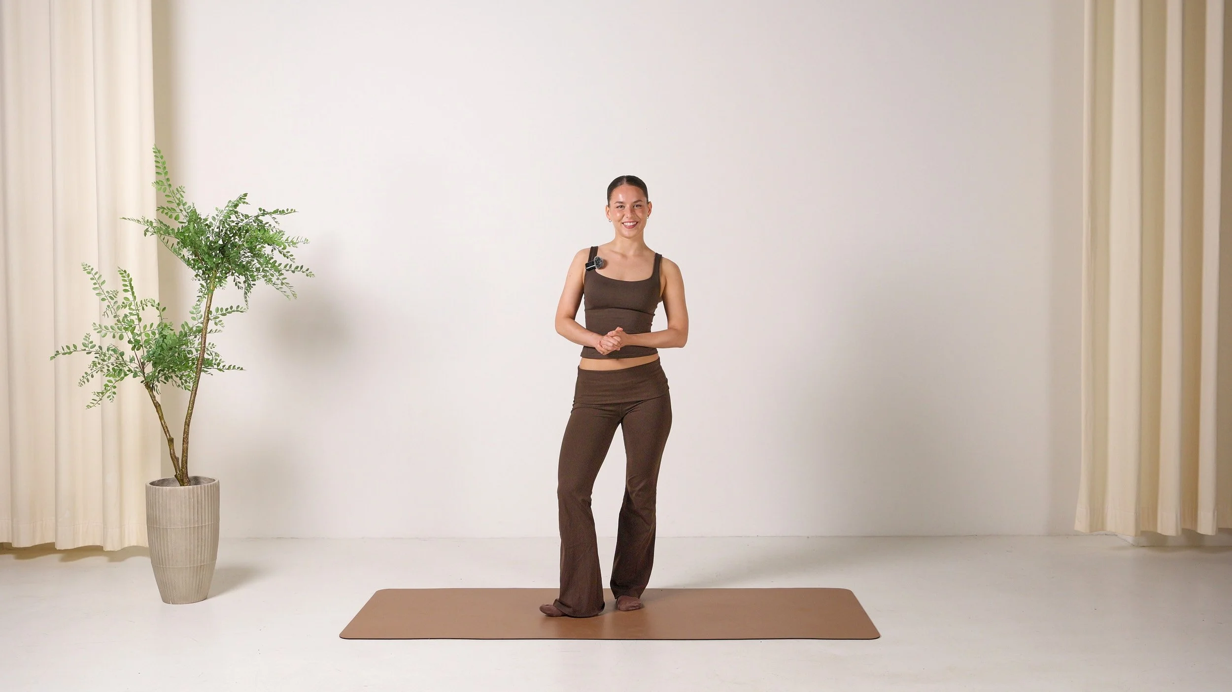 A woman standing on a pilates mat in a room with a potted plant, beige curtains, and a plain wall, smiling at the camera.