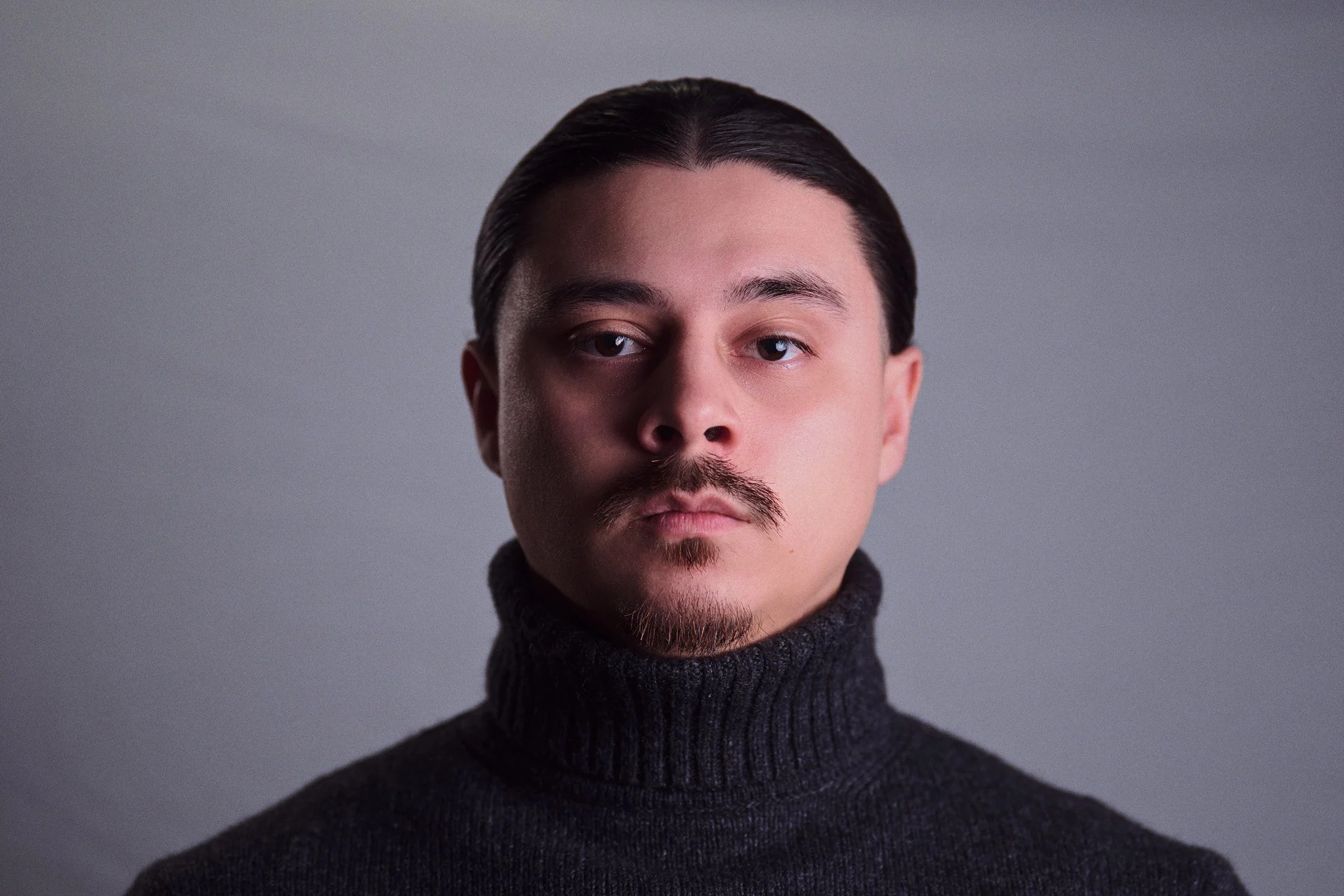 Close-up portrait of a young man with dark hair, styled back, and a mustache and goatee, wearing a black turtleneck sweater against a gray background.