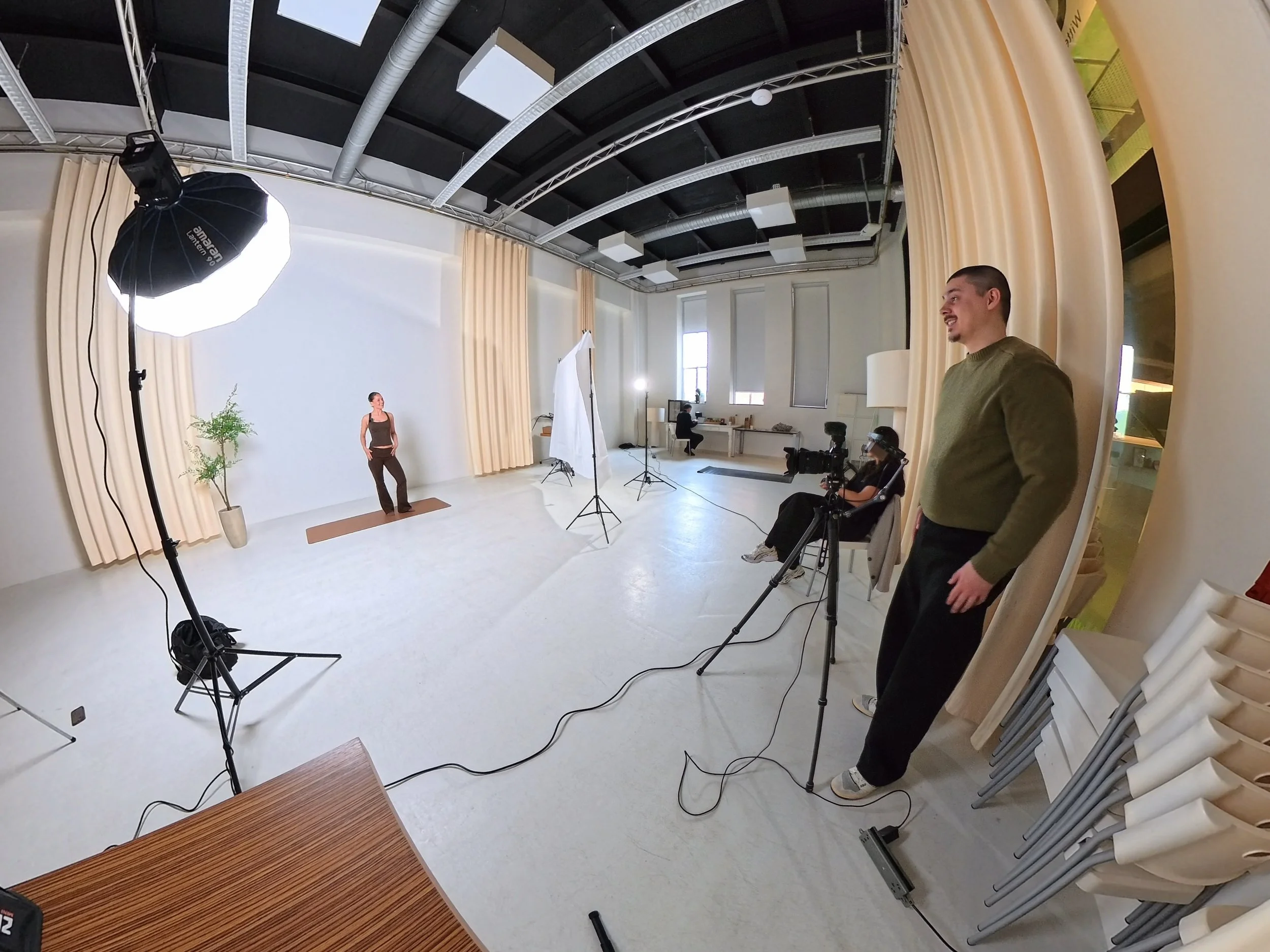 Film studio with a woman posing for a Video, a man standing near curtains, professional lighting equipment, chairs stacked against the wall, and a person working at a desk in the background.