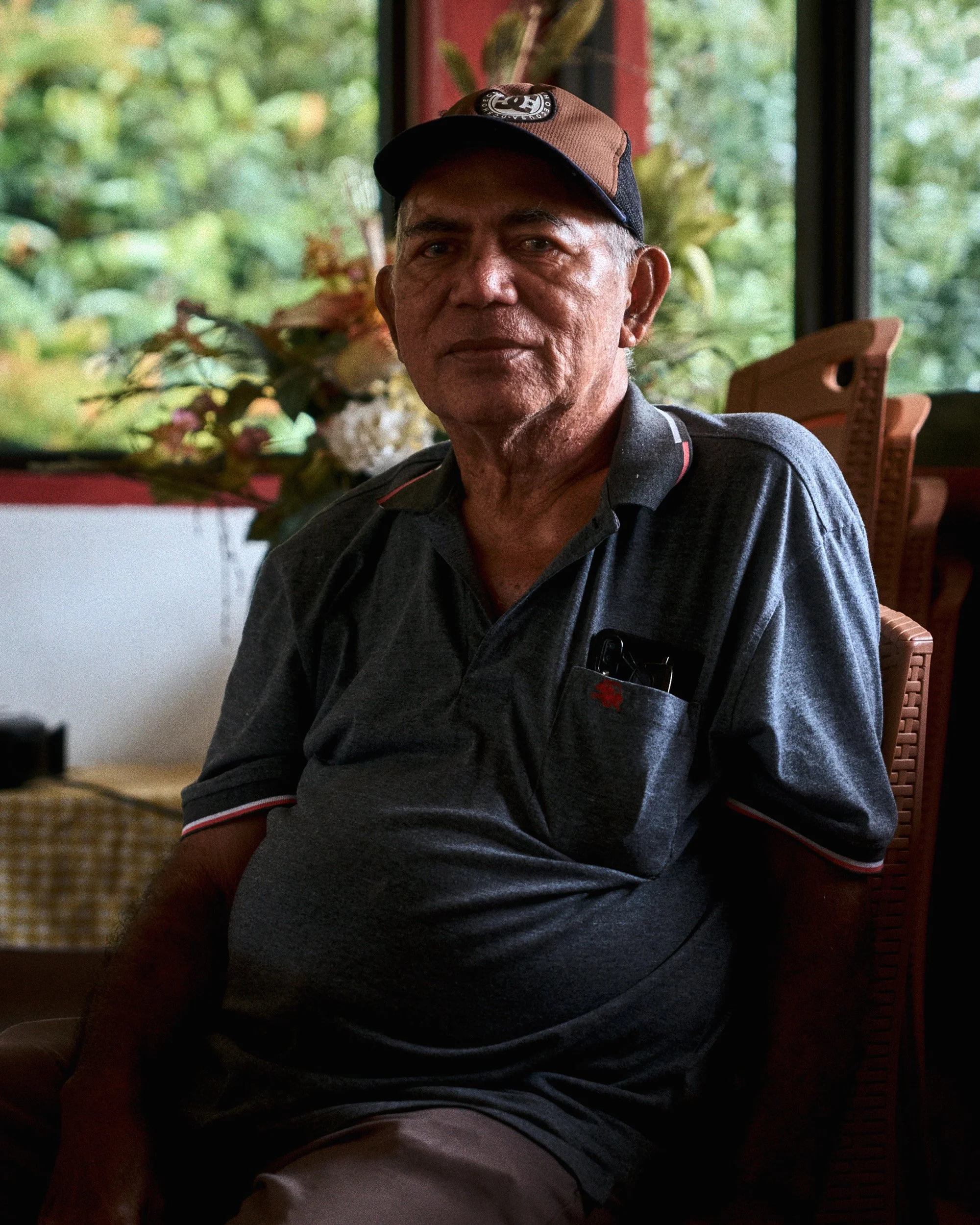 A middle-aged man with short hair and tanned skin, wearing a dark cap with a logo and a gray polo shirt, sitting indoors with a background of green foliage outside the window.