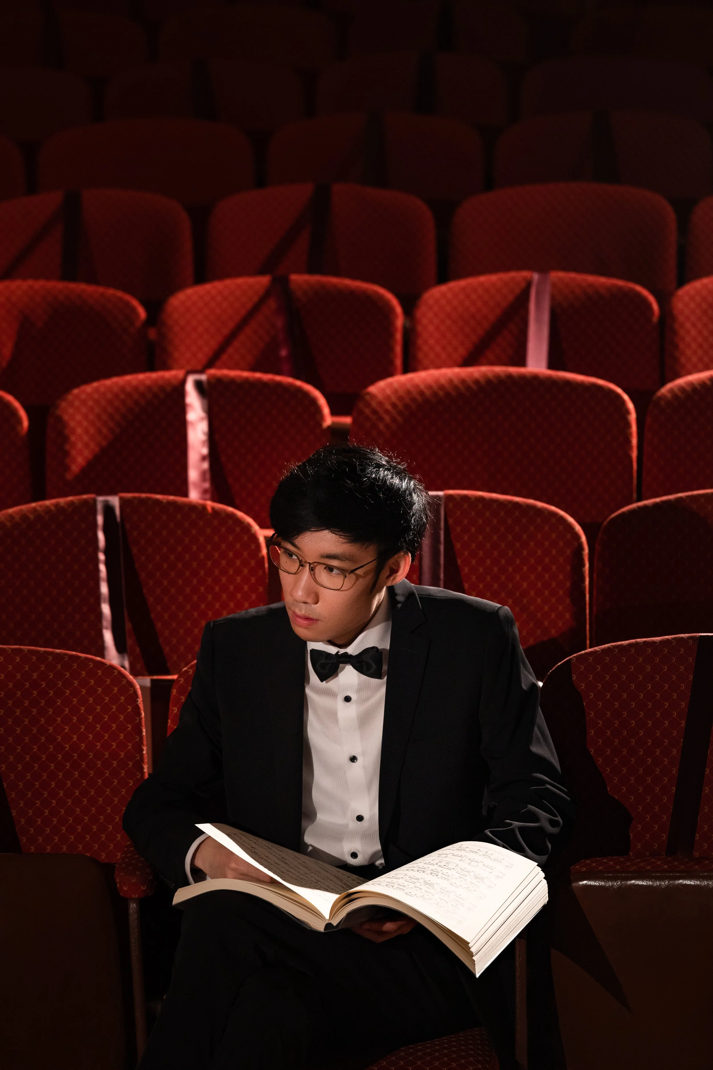 A young man dressed in a tuxedo with a bow tie, sitting in an empty theatre with red cushioned seats, reading a large open book or music score.