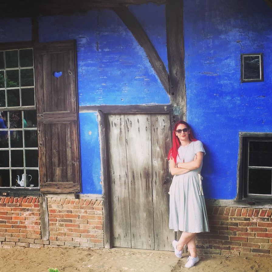 A woman with red hair, sunglasses, and a gray dress stands with arms crossed leaning against a rustic blue wall with windows and a wooden door.