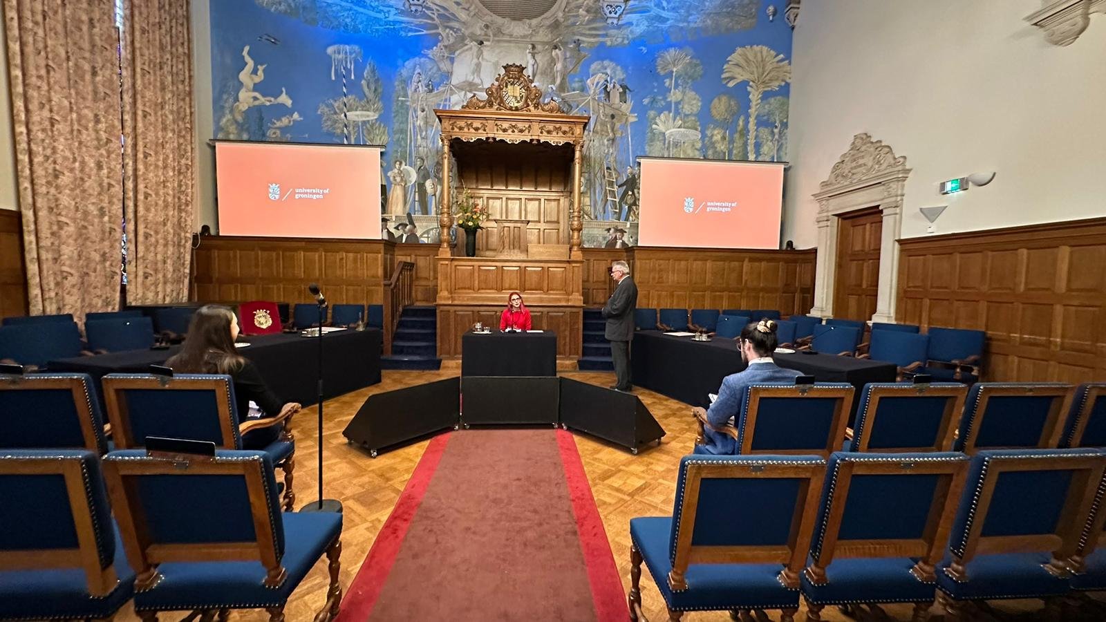 A spacious conference room with wooden paneling, a raised stage with a wooden throne-like seat, two large screens displaying the University of Groningen logo, and people sitting and standing during a formal event or presentation.