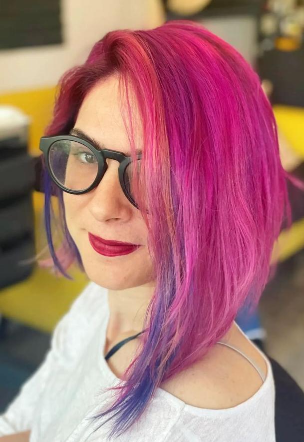 Woman with pink and purple hair, wearing glasses and red lipstick, smiling slightly in an indoor setting.