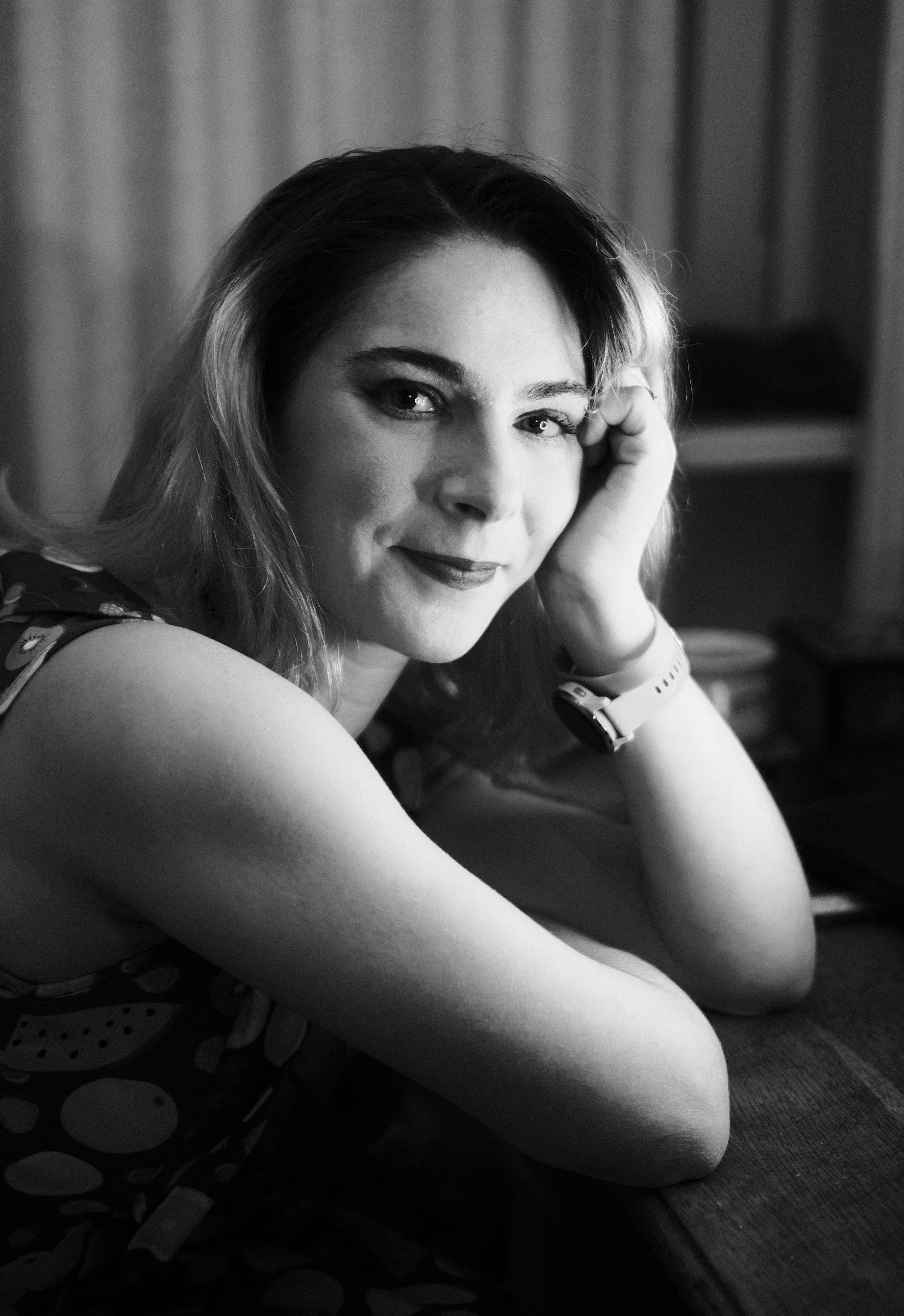 A black and white portrait of a woman with light skin, wavy hair, and expressive eyes, resting her face on her hand with a slight smile.