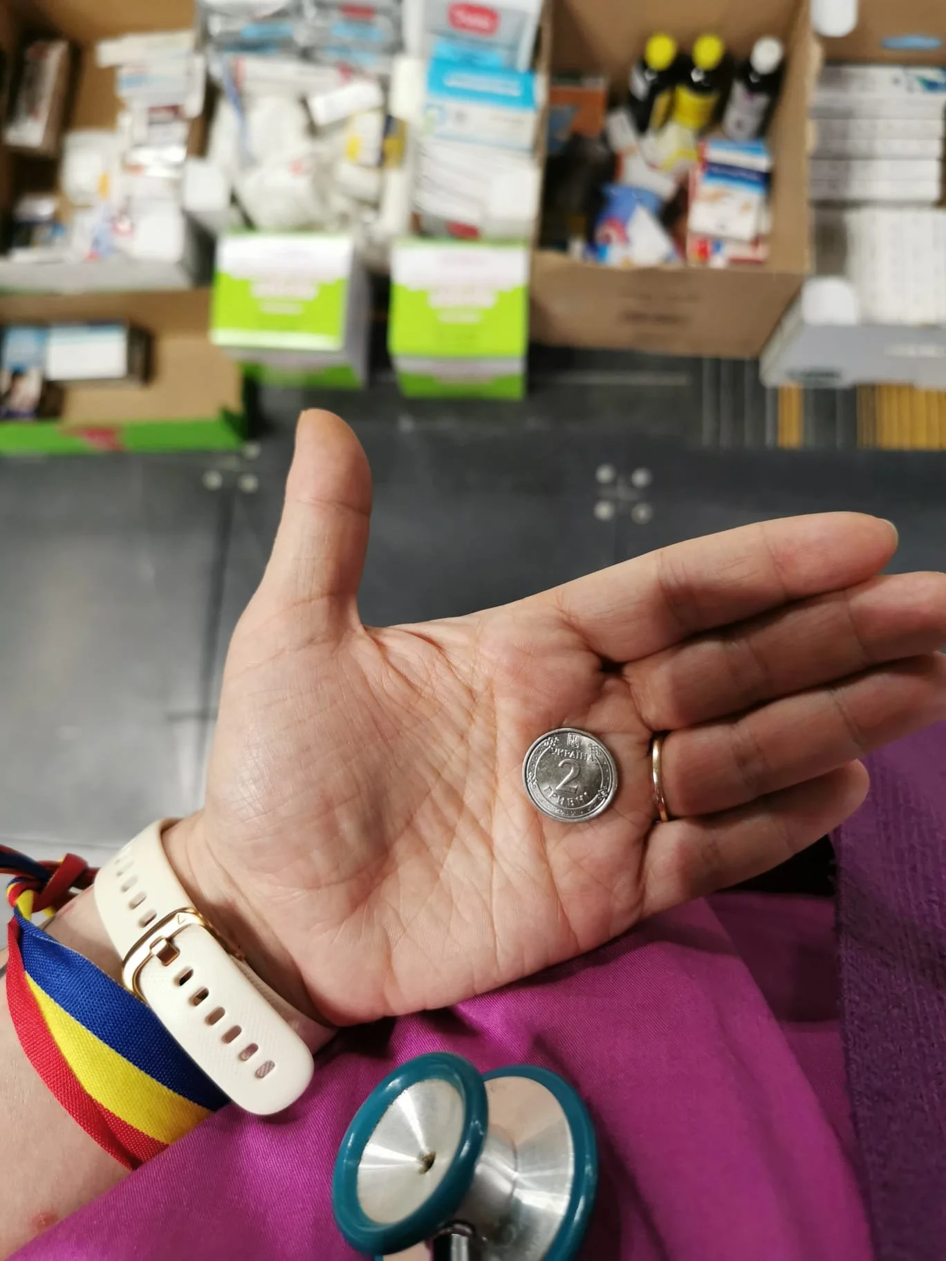 A person holding a 2 peso coin in their left hand, with a stethoscope in their lap, inside a store with medical supplies on shelves in the background.