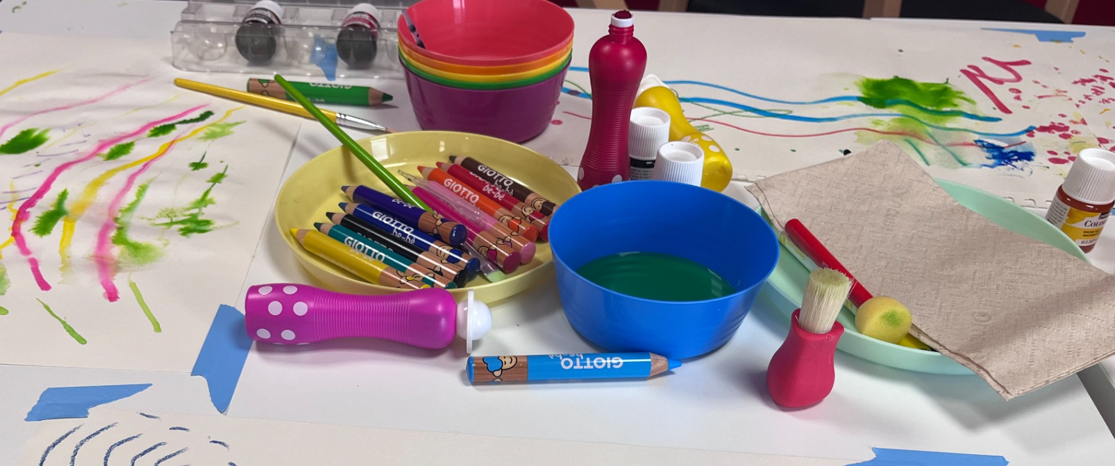 Table d'arts plastiques avec des pinceaux, peintures, feutres, crayon, feuilles, et autres fournitures pour peinture, avec des dessins colorés en arrière-plan.
