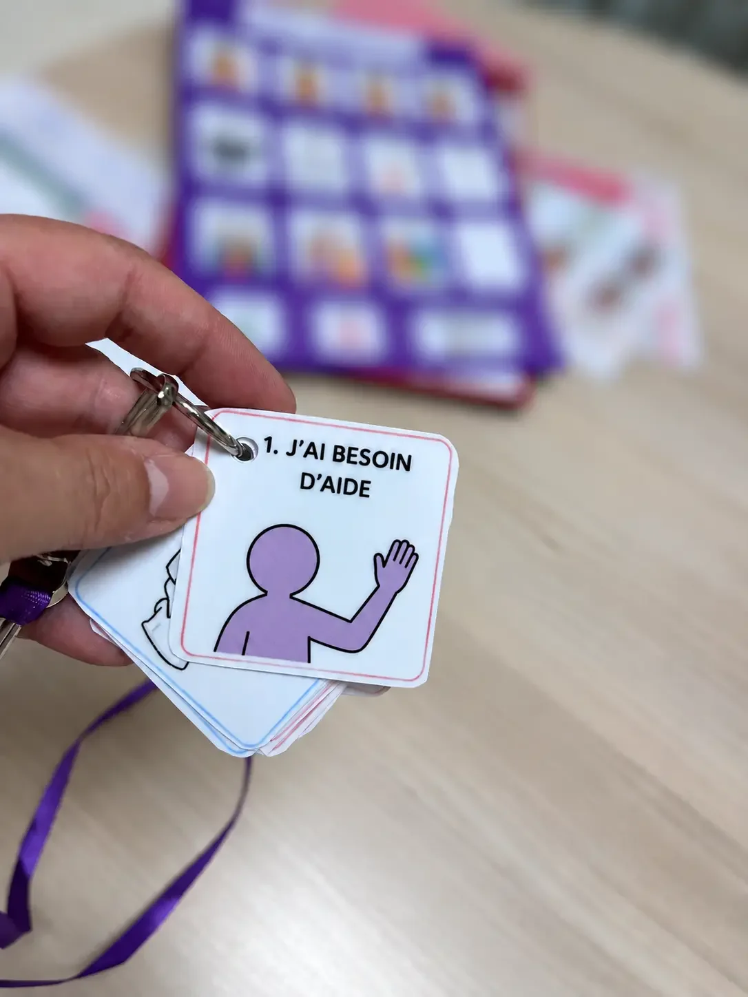 Une main tient un pictogramme avec le texte "J'AI BESOIN D'AIDE" et une illustration d'une personne faisant un geste de la main. En arrière-plan, il y a une planche de pictogrammes de CAA sur une table.
