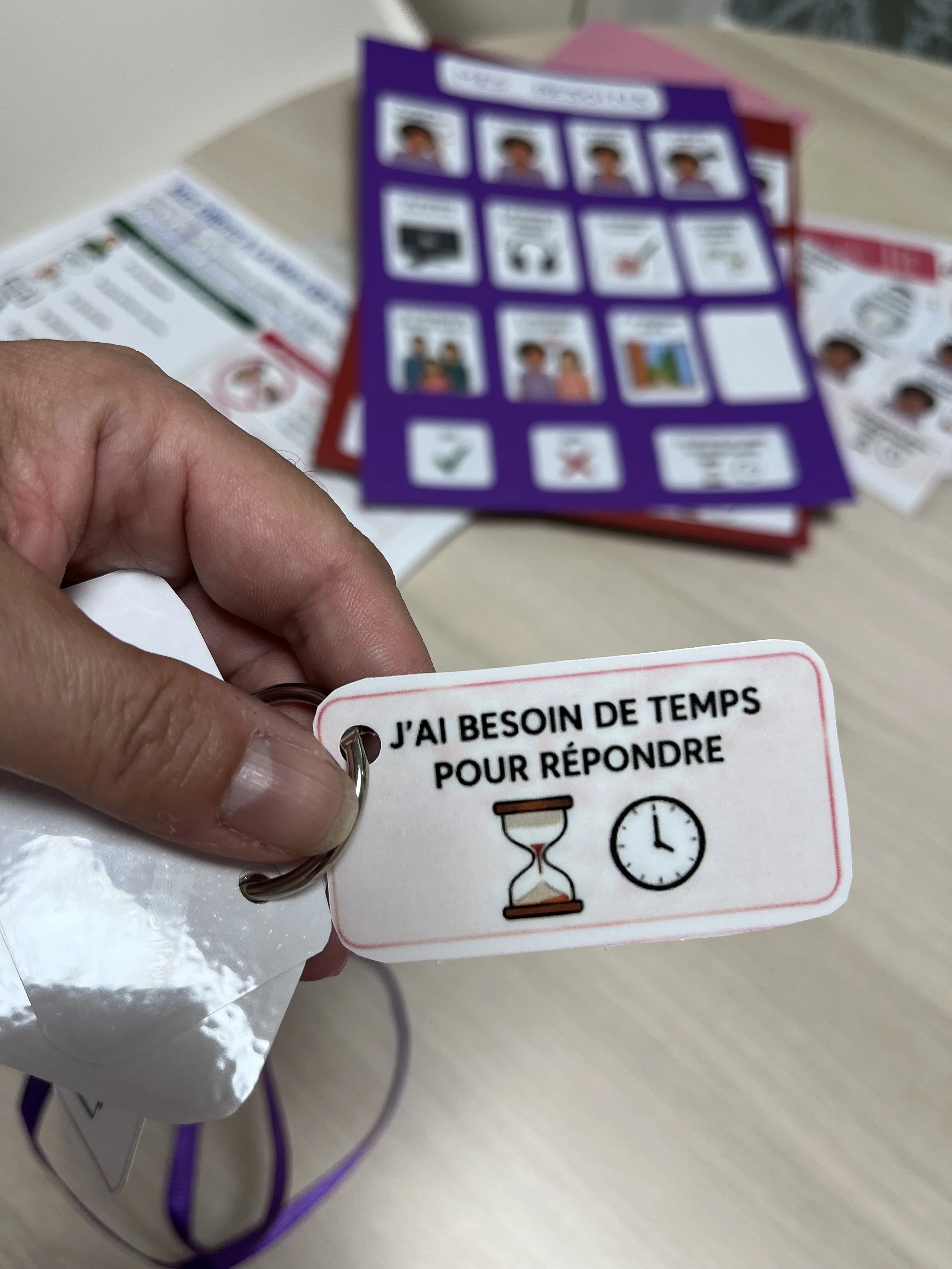 Une main montre un dispositif de CAA avec un badge illustré, affichant des pictogrammes de temps et de réponse, sur une table avec des brochures en arrière-plan.