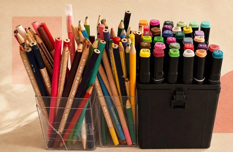 Une boîte contenant des crayons de couleur et des marqueurs de différentes couleurs sur une surface beige.
