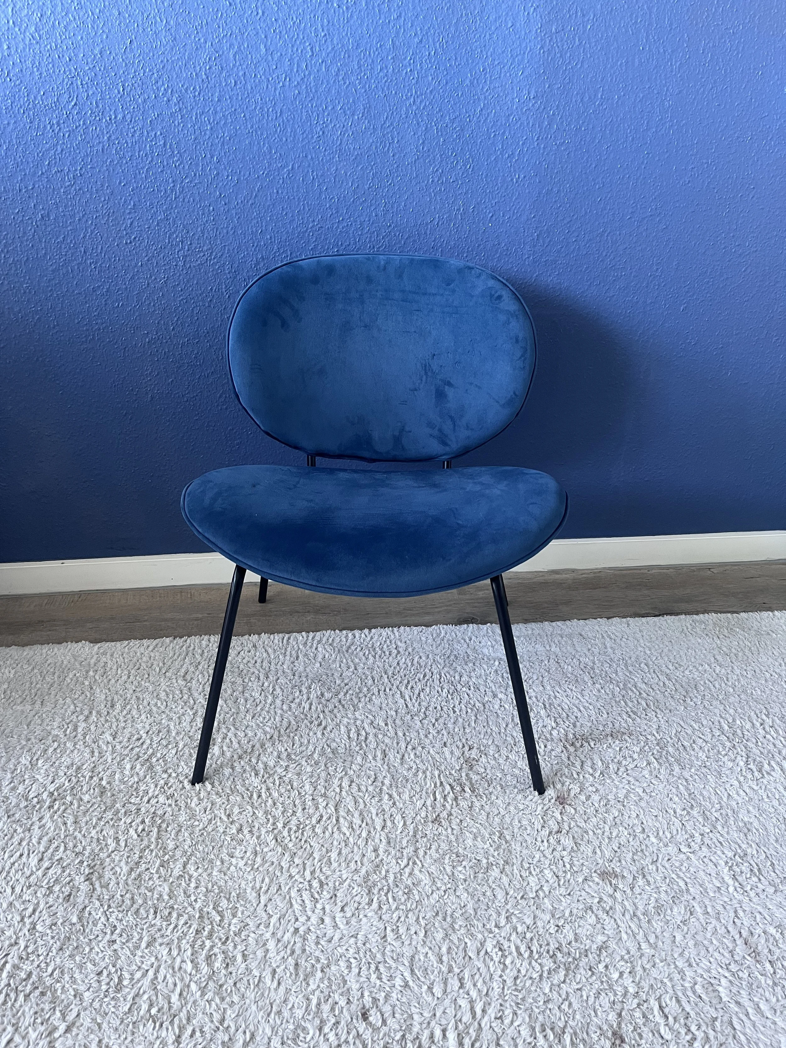 Une chaise bleue en velours placée sur un tapis blanc, contre un mur bleu.