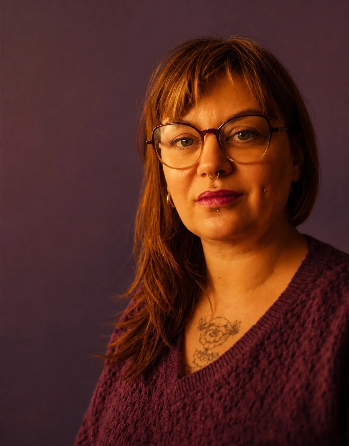 Portrait d'une femme aux cheveux bruns, portant des lunettes, un tatouage floral sur le décolleté, vêtue d'un pull violet, avec un fond violet.