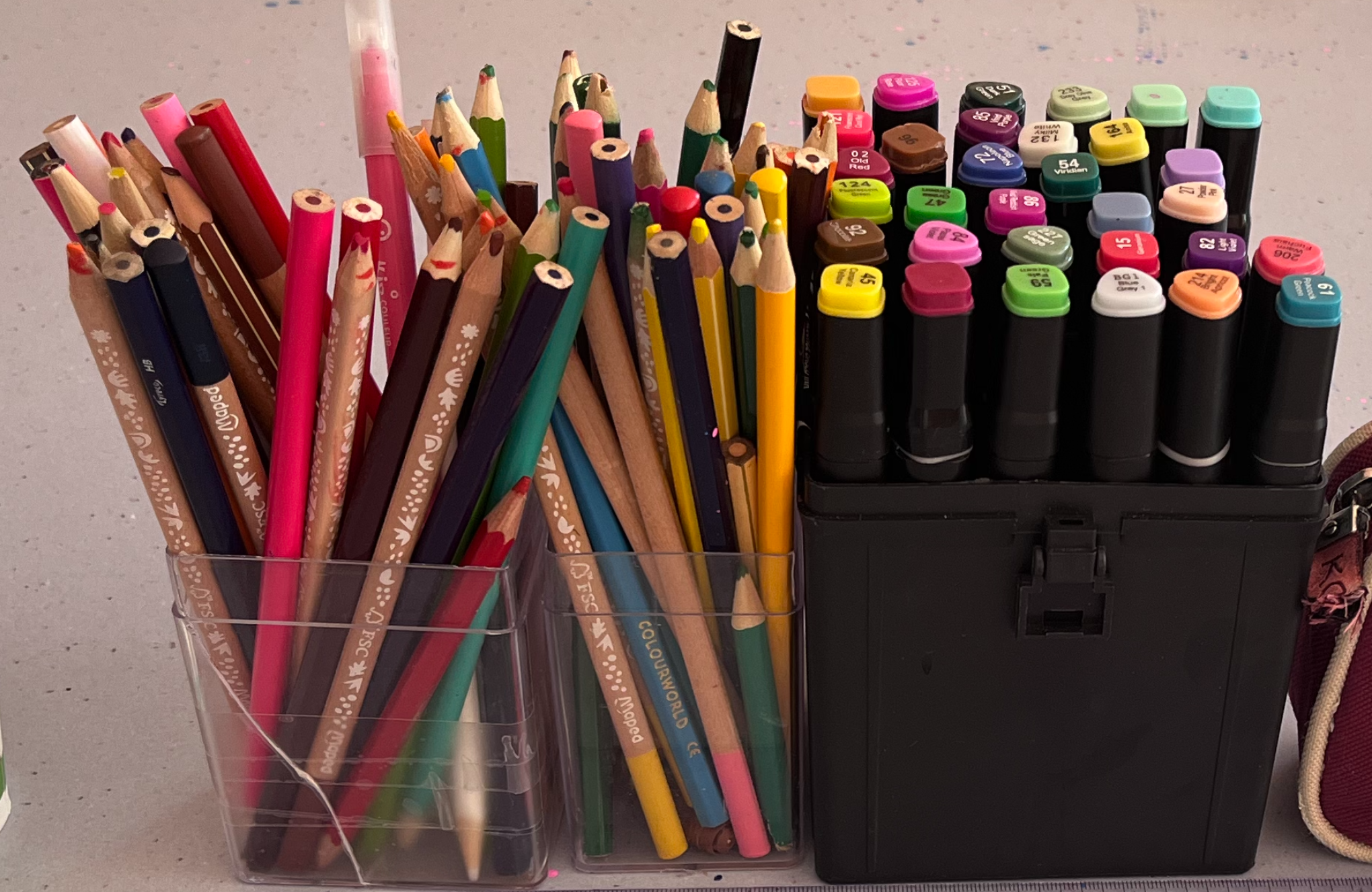 Un ensemble de crayons de couleur, feutres, et autres outils d'écriture dans des boîtes en plastique, posés sur une surface grise.