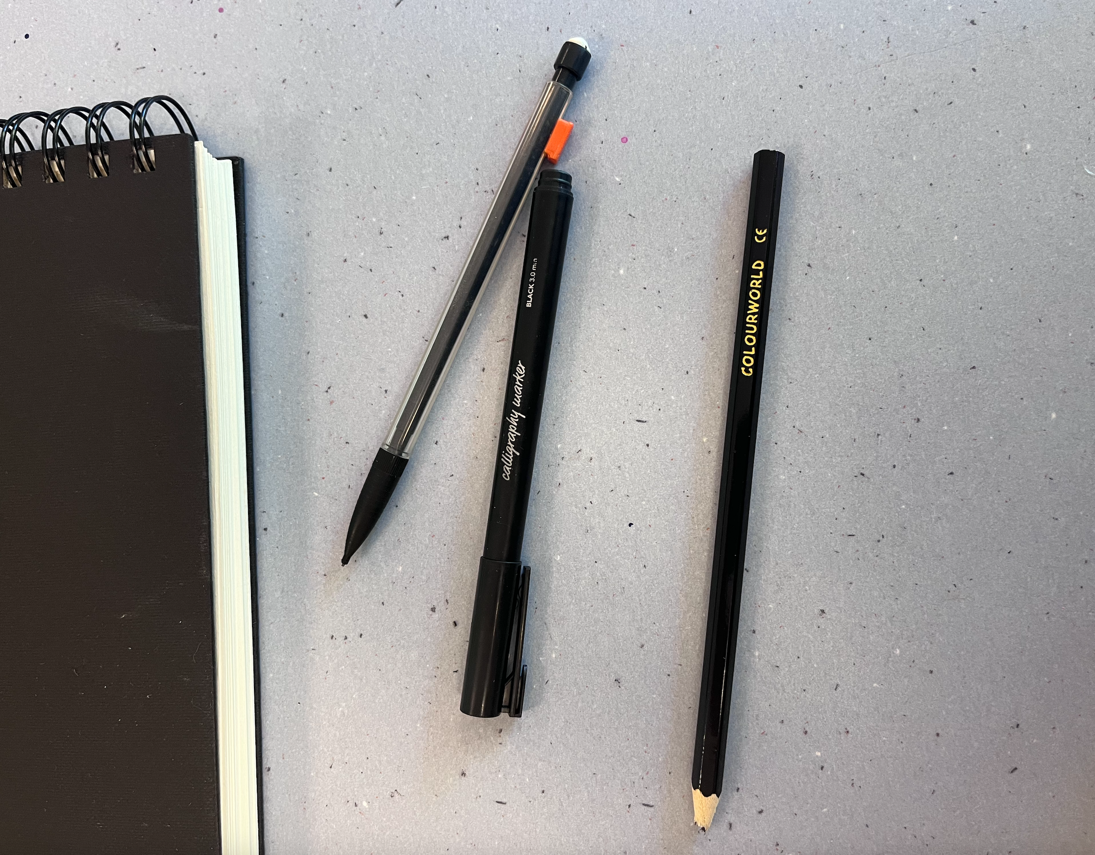 Un carnet noir, deux stylos noirs et un crayon sont posés sur une surface grise.