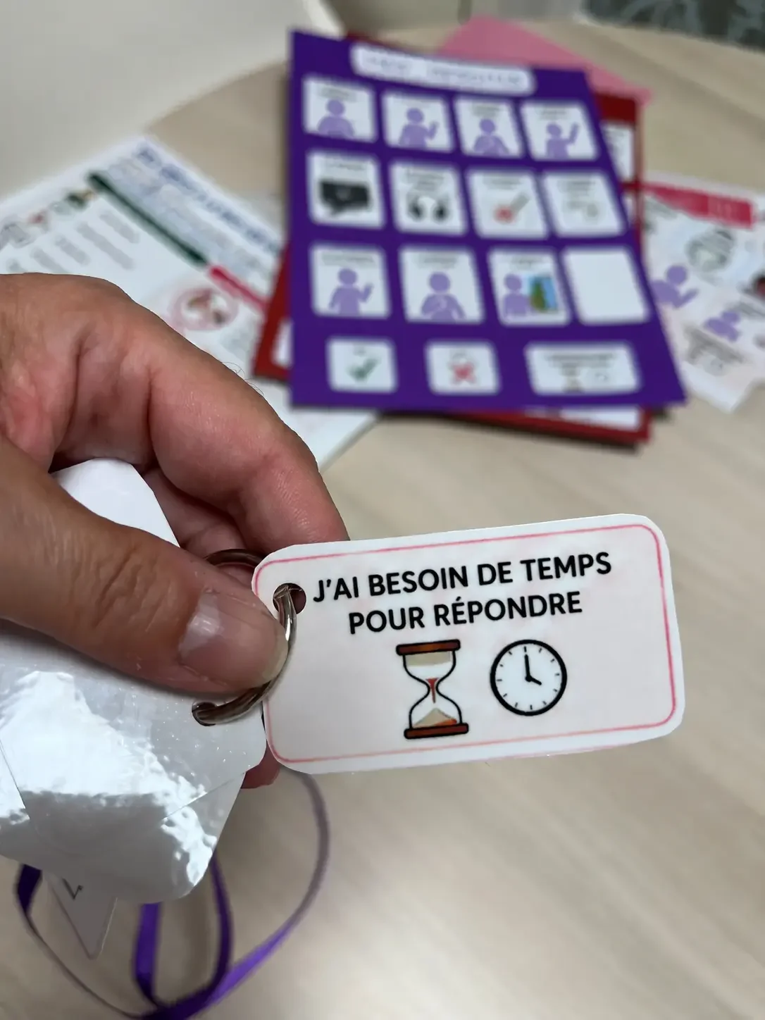 Une personne tient un pictogramme imprimmé avec le texte "J'AI BESOIN DE TEMPS POUR RÉPONDRE" accompagnée d'une horloge et d'un sablier, avec des documents de CAA flous en arrière-plan.
