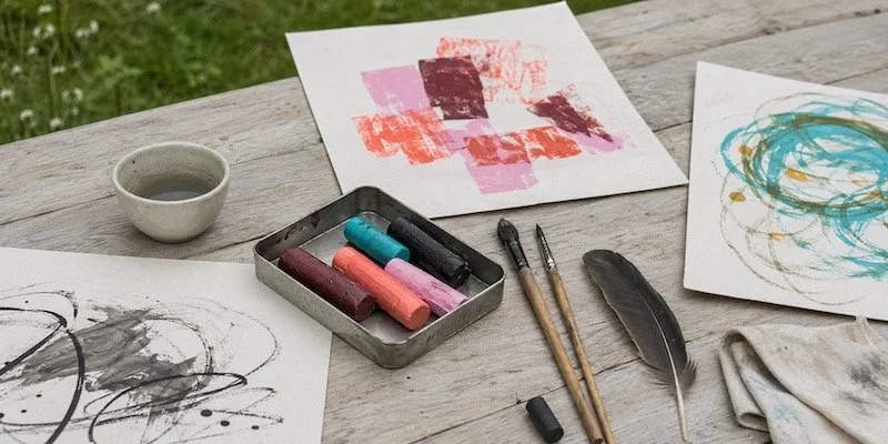 Table extérieure en bois avec des œuvres d'art abstraites à la craie, des pinceaux, un petit bol, une plume, une éponge et un chiffon.