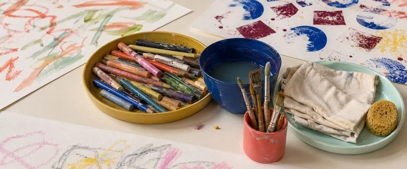 Table avec fournitures d'art, comprenant des bâtons de pastel, un pot d'eau, un récipient avec des pinceaux, des chiffons, et une éponge, ainsi que des œuvres d'art en cours de création.